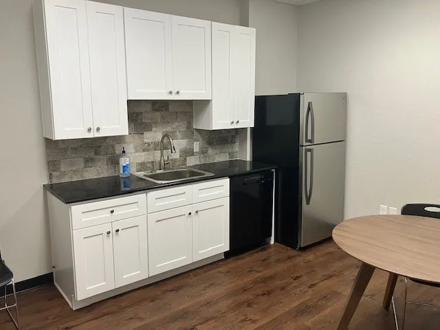 Compact kitchen area featuring white shaker-style cabinetry, a stainless steel sink with a gooseneck faucet, black countertops, and a stacked stone-look backsplash