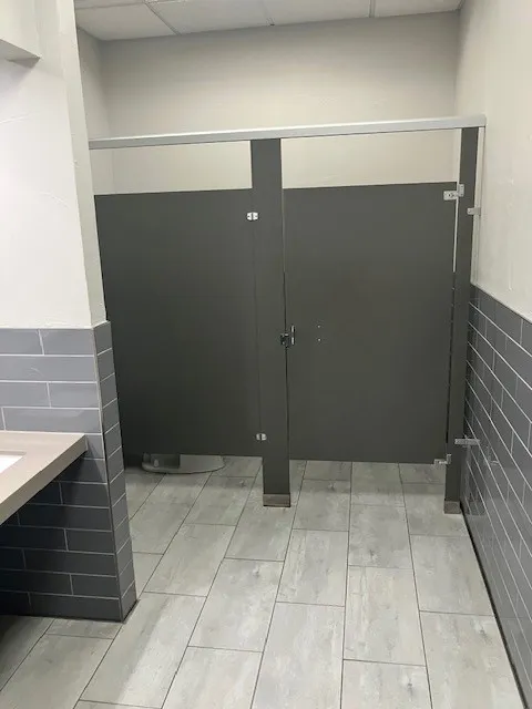 Restroom space featuring gray tile walls, wood-finish tile flooring, and dark privacy partitions