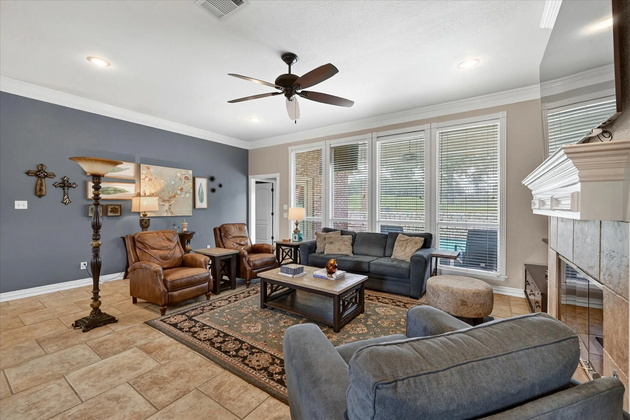 Living area featuring large format tile flooring, a fireplace with a decorative mantel, white crown molding, and a ceiling fan with integrated lighting
