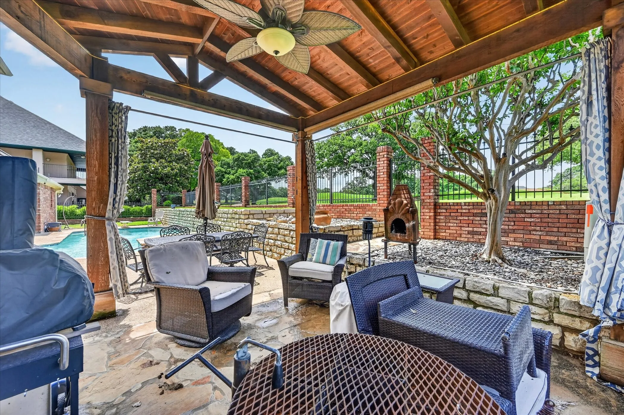 Expansive outdoor patio featuring a wood-beamed ceiling with a ceiling fan, flagstone flooring, a stone retaining wall, and a brick-and-wrought iron fence