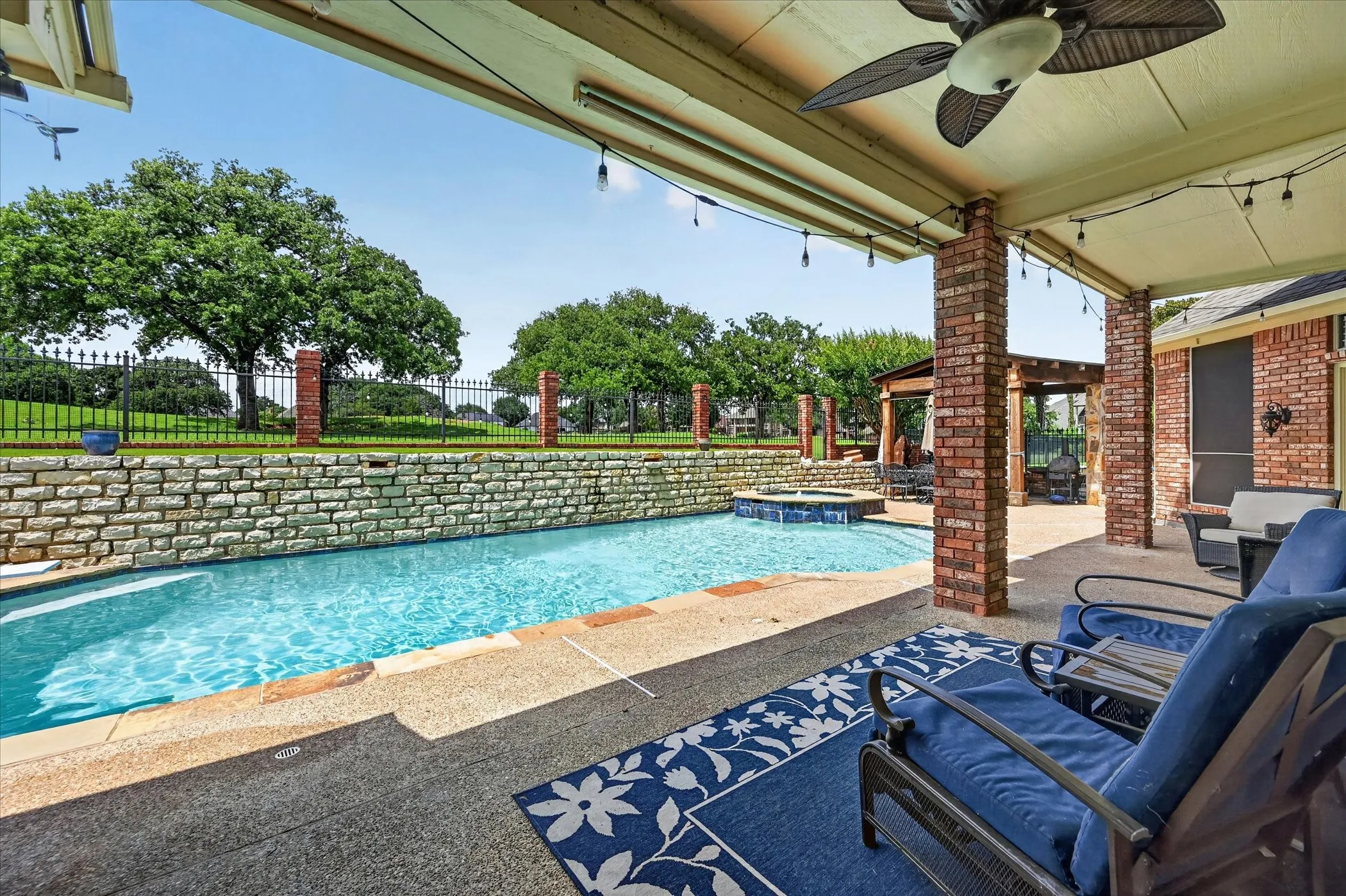 Covered patio with a ceiling fan and string lights, overlooking a swimming pool with an integrated spa, stone retaining wall, and a wrought iron fence
