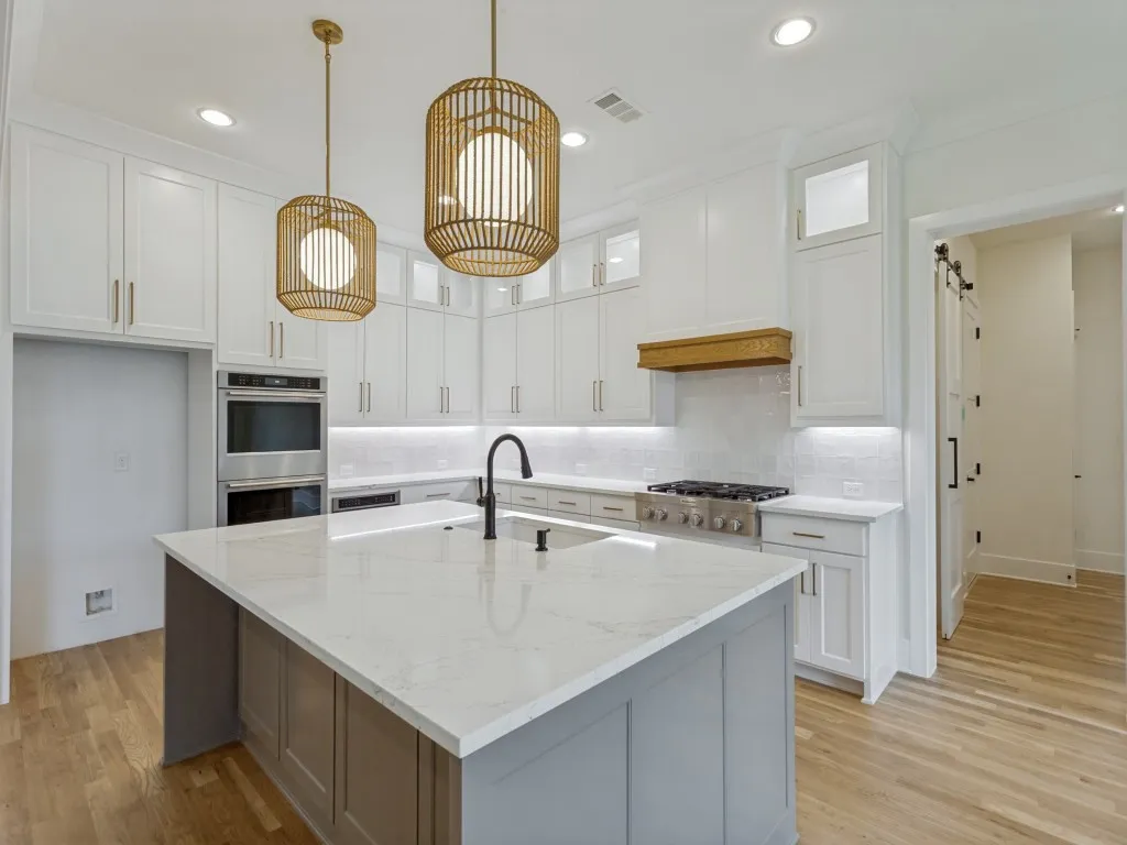 Modern kitchen featuring a large central island with a light-toned countertop, white cabinetry with gold-toned hardware, stainless steel appliances, and wood-finish flooring