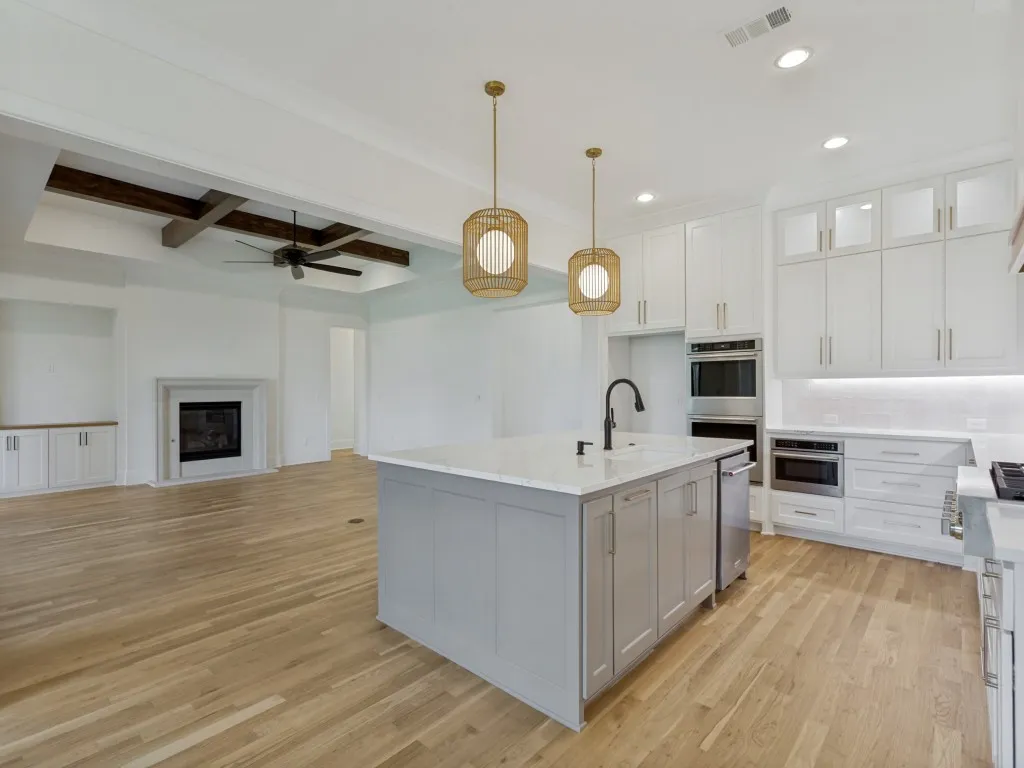 Open-concept kitchen featuring a large island with a countertop sink and matte black faucet, white shaker cabinetry with gold-toned hardware, and stainless steel built-in appliances