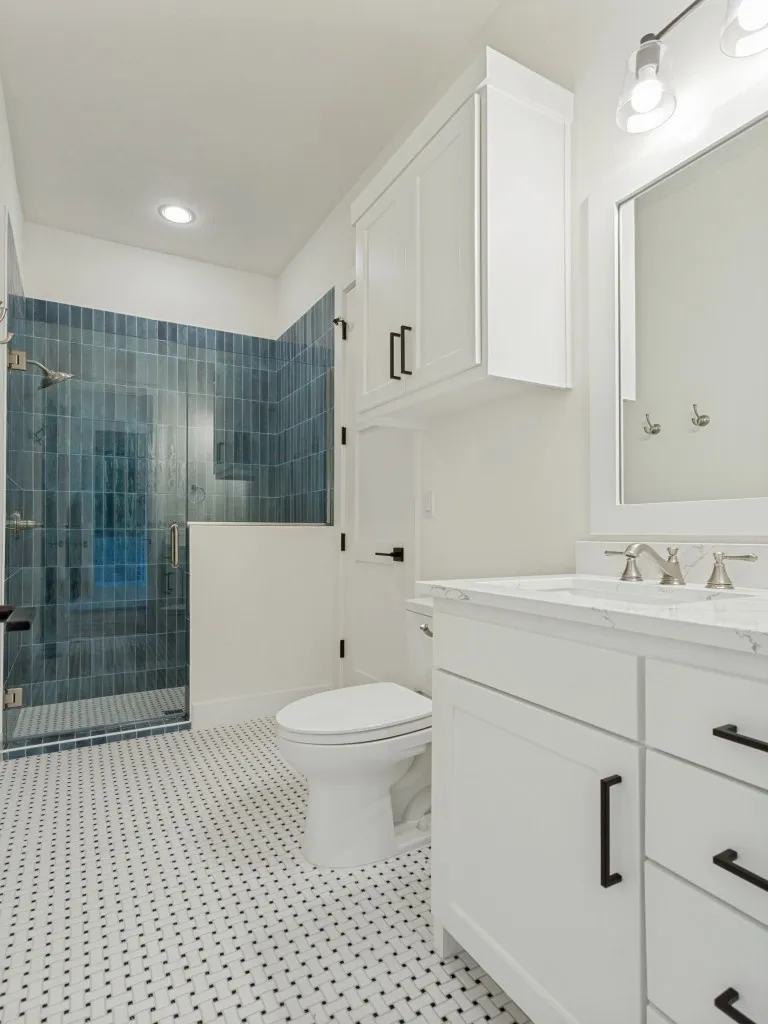 Bathroom featuring a glass-enclosed shower with blue vertical tile, white vanity with a light-toned countertop, white cabinetry with black hardware, and mosaic tile flooring