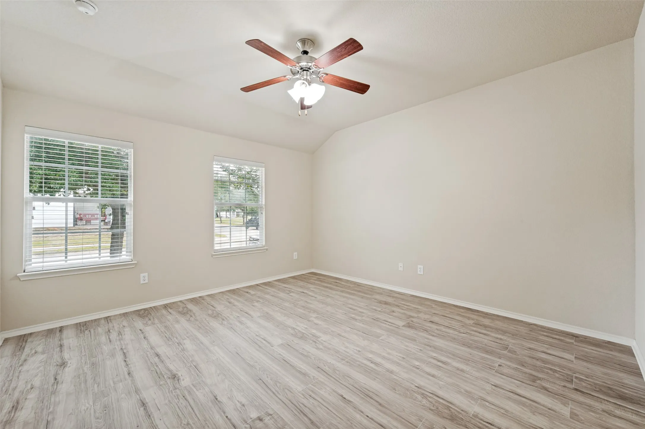 Spacious room featuring light-toned walls, wood-finish flooring, two windows with blinds, a ceiling fan with light fixture, and a vaulted ceiling