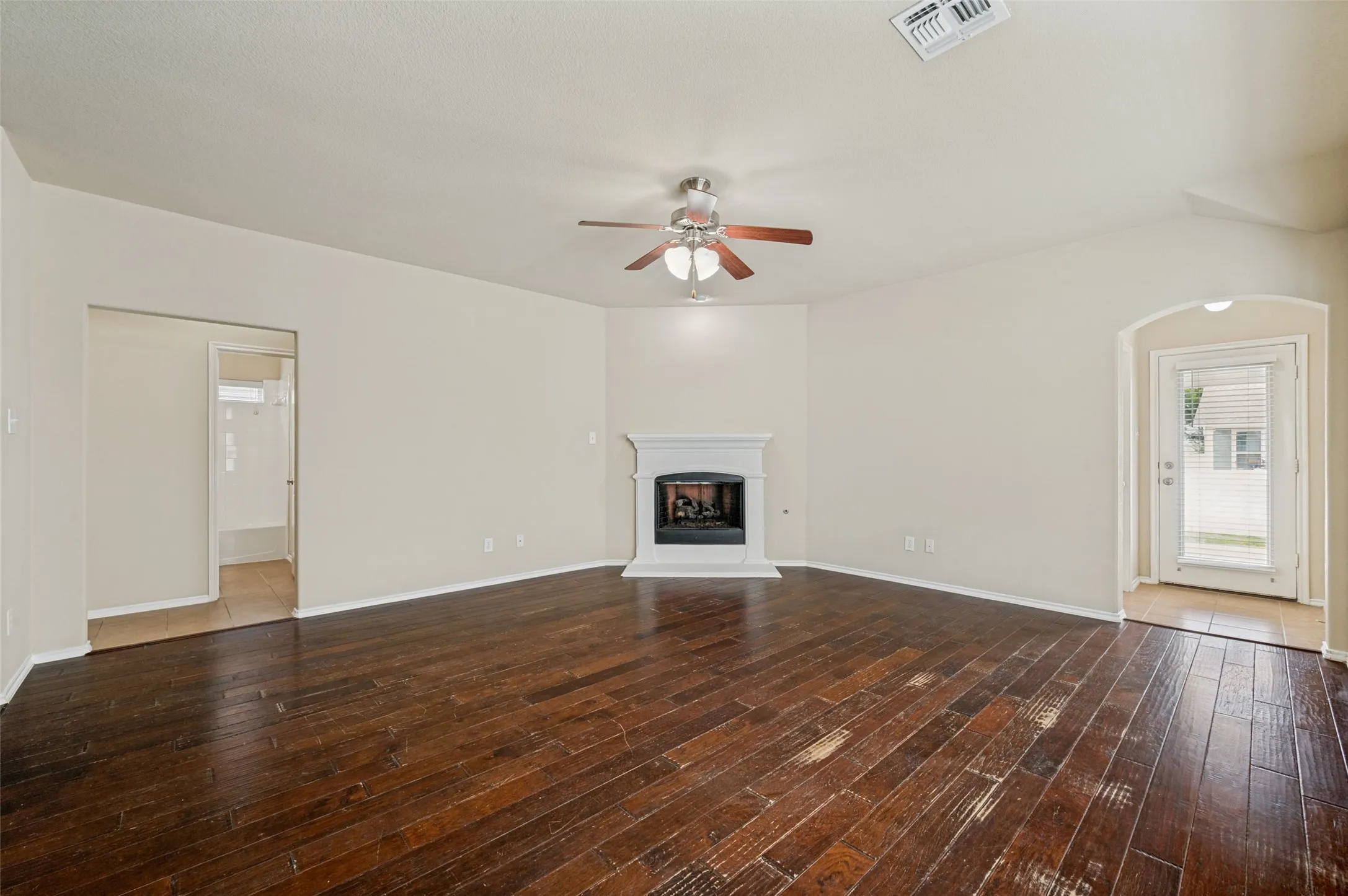 Spacious living area featuring wood-finish flooring, a white mantel fireplace, a ceiling fan with integrated lighting, and an arched entryway