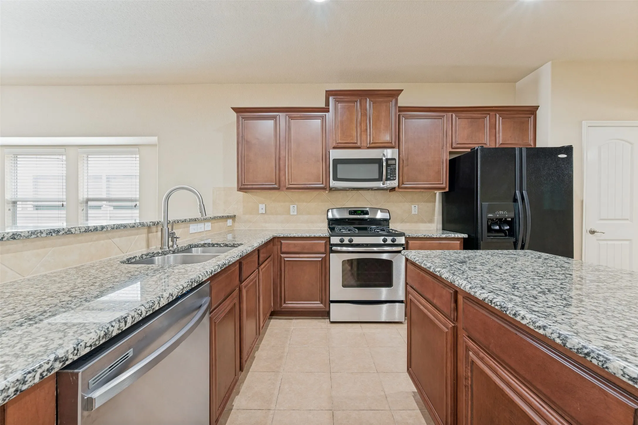 Kitchen featuring granite countertops, wood cabinetry, stainless steel appliances, a dual-basin sink with a gooseneck faucet, and tile flooring