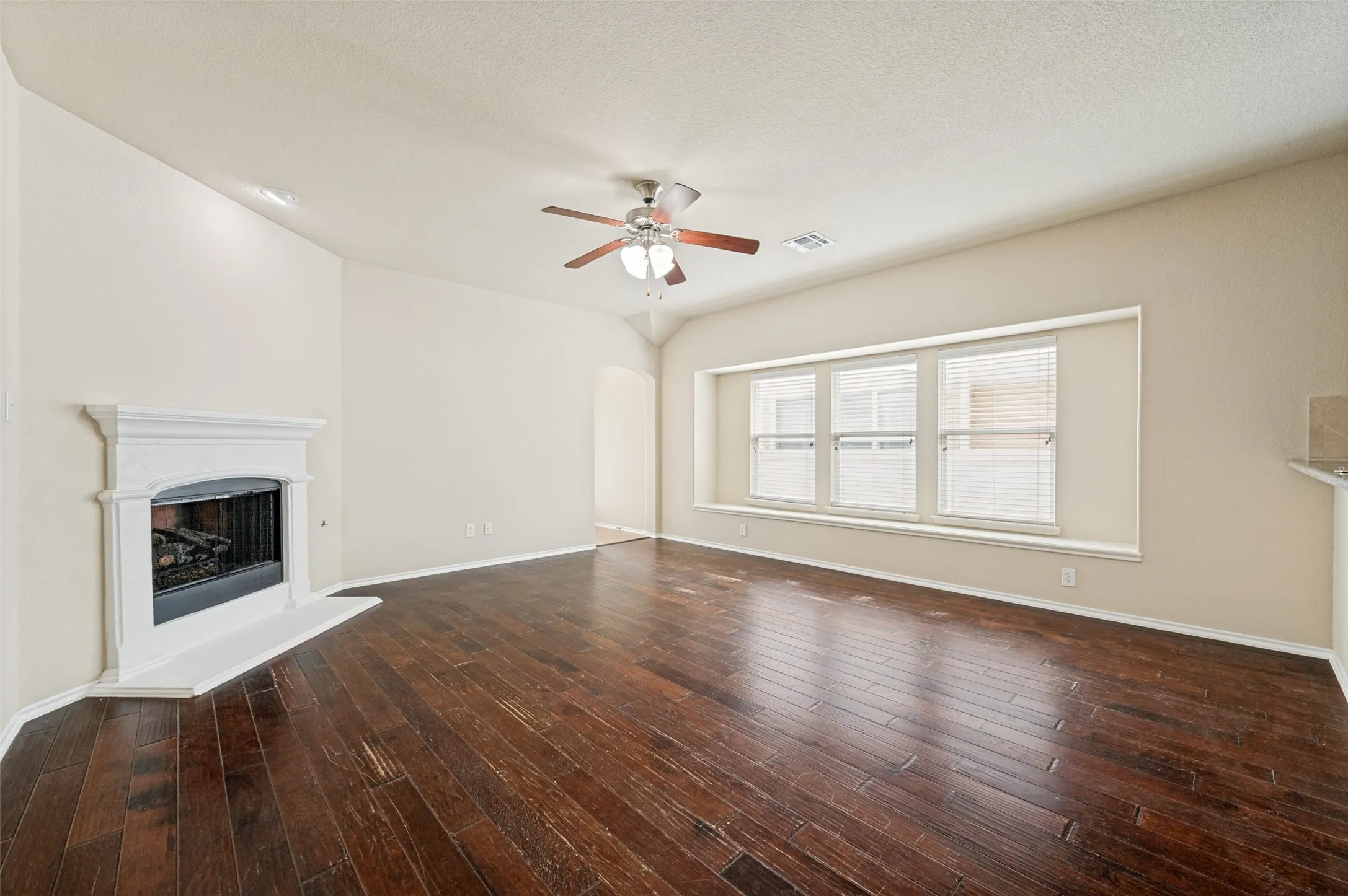 Spacious room featuring rich wood-finish flooring, a white mantel fireplace, three large windows with blinds, a built-in window seat, and a ceiling fan