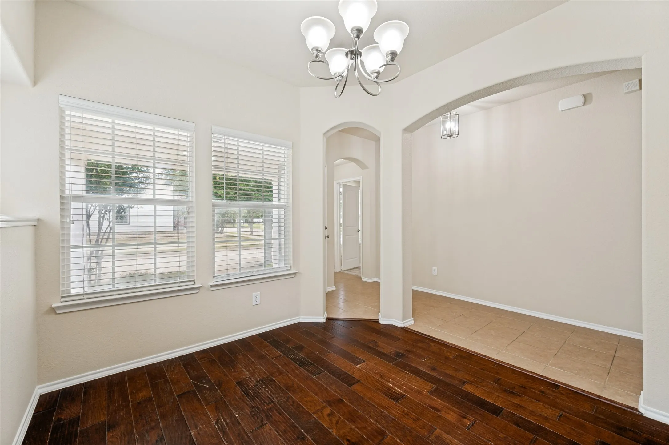 Interior space featuring rich wood-finish flooring, two large windows with blinds, a multi-bulb chandelier, and an arched entryway leading to a tiled area with a decorative pendant light