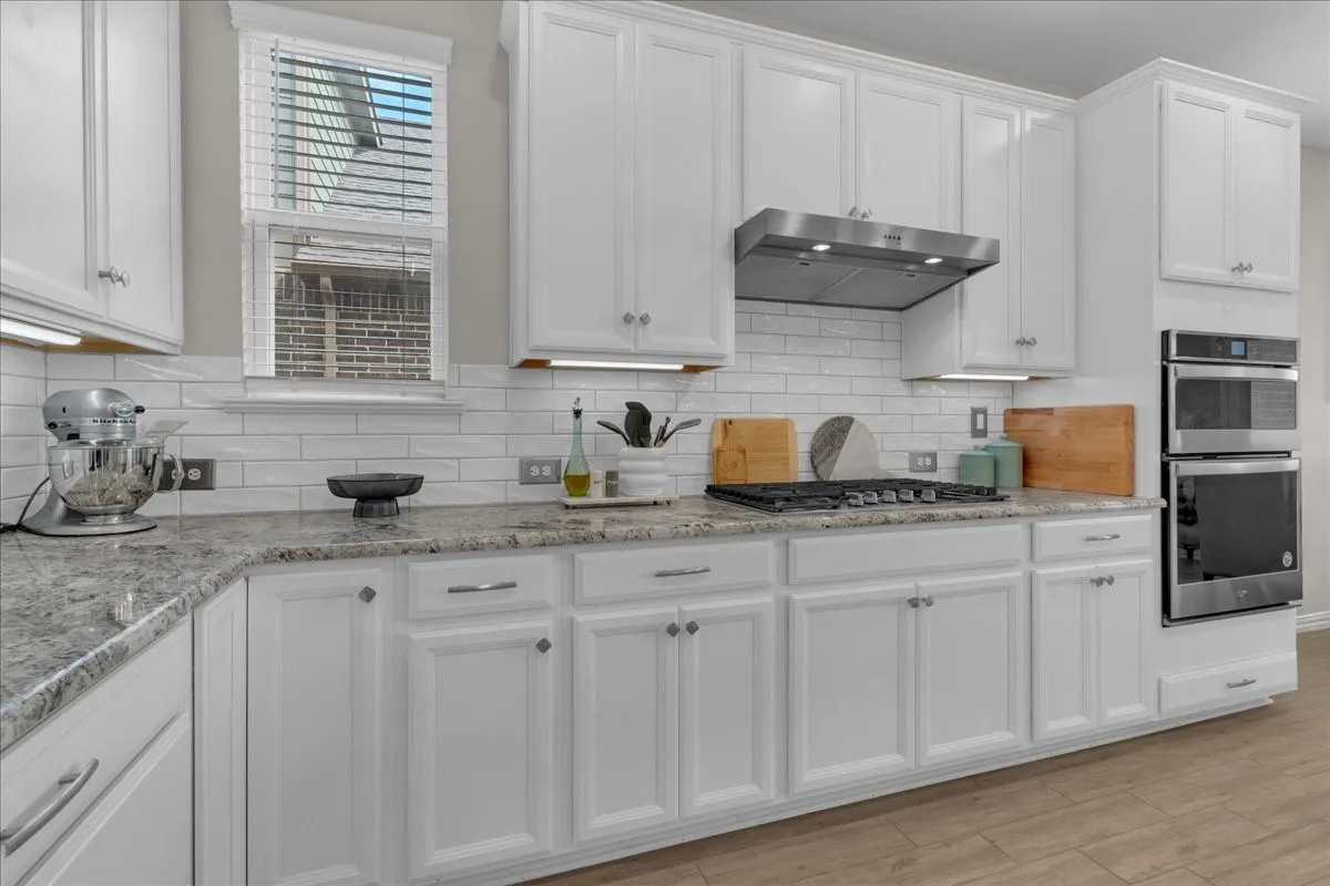 Kitchen featuring white paneled cabinetry, light-colored speckled countertops, subway tile backsplash, stainless steel gas range, and built-in double ovens