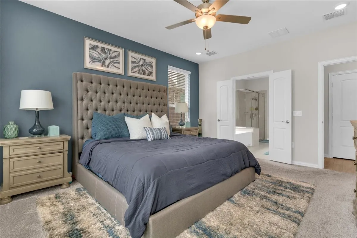 Carpeted bedroom featuring a ceiling fan with integrated lighting, a contrasting accent wall, and a large doorway opening to a bathroom with a shower and soaking tub