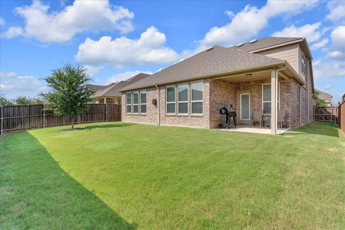 Expansive backyard featuring a manicured lawn, a mature tree, a full privacy fence, a covered patio, and a brick exterior