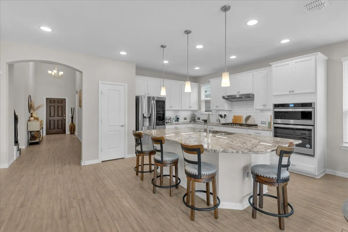 Spacious kitchen featuring an expansive center island with seating, pendant lighting, white cabinetry, integrated stainless steel appliances, and wood-finish flooring
