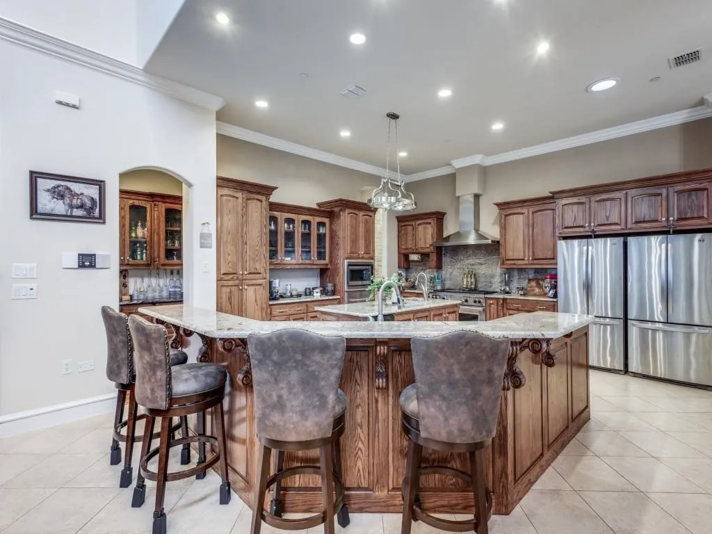 Spacious kitchen featuring an expansive multi-level island with seating, natural wood cabinetry, light-toned tile flooring, and stainless steel appliances, including a double refrigerator and range hood