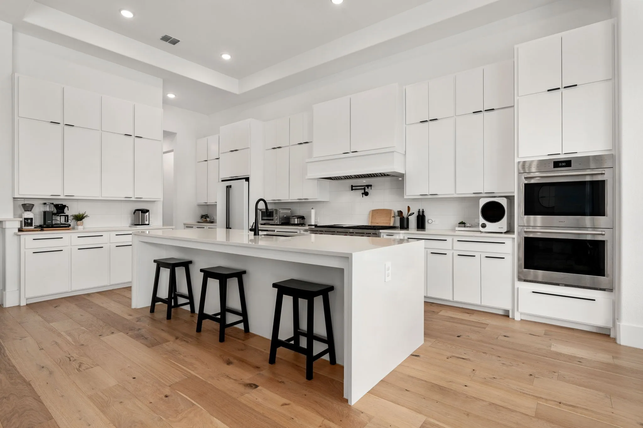 Expansive kitchen featuring an oversized island with countertop seating, white shaker cabinetry extending to the ceiling, stainless steel double wall ovens, wood-finish flooring, and recessed lighting