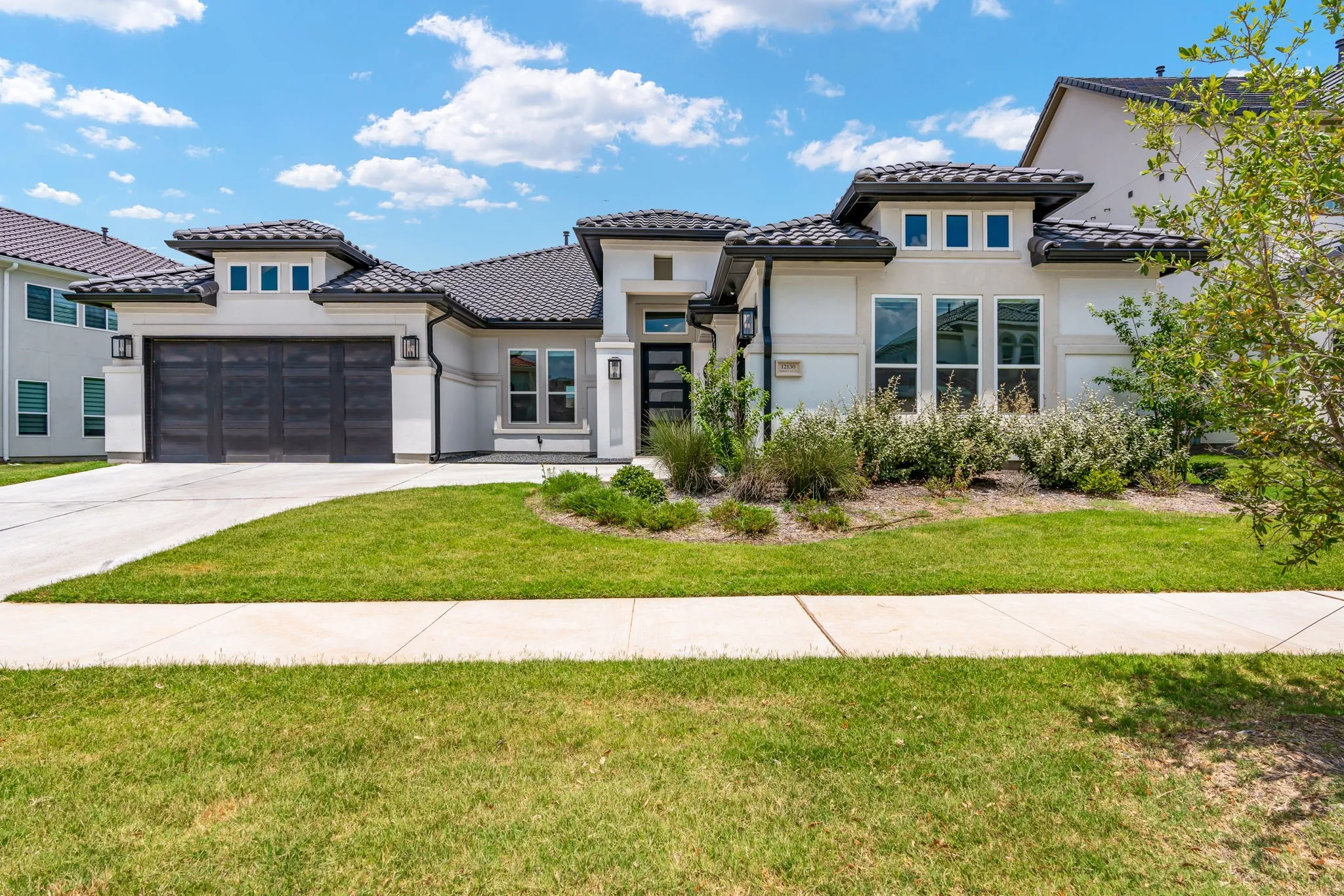 Contemporary stucco exterior featuring a dark tile roof, a two-car garage with a textured door, a minimalist front door with frosted glass panels, and multiple large windows with dark trim