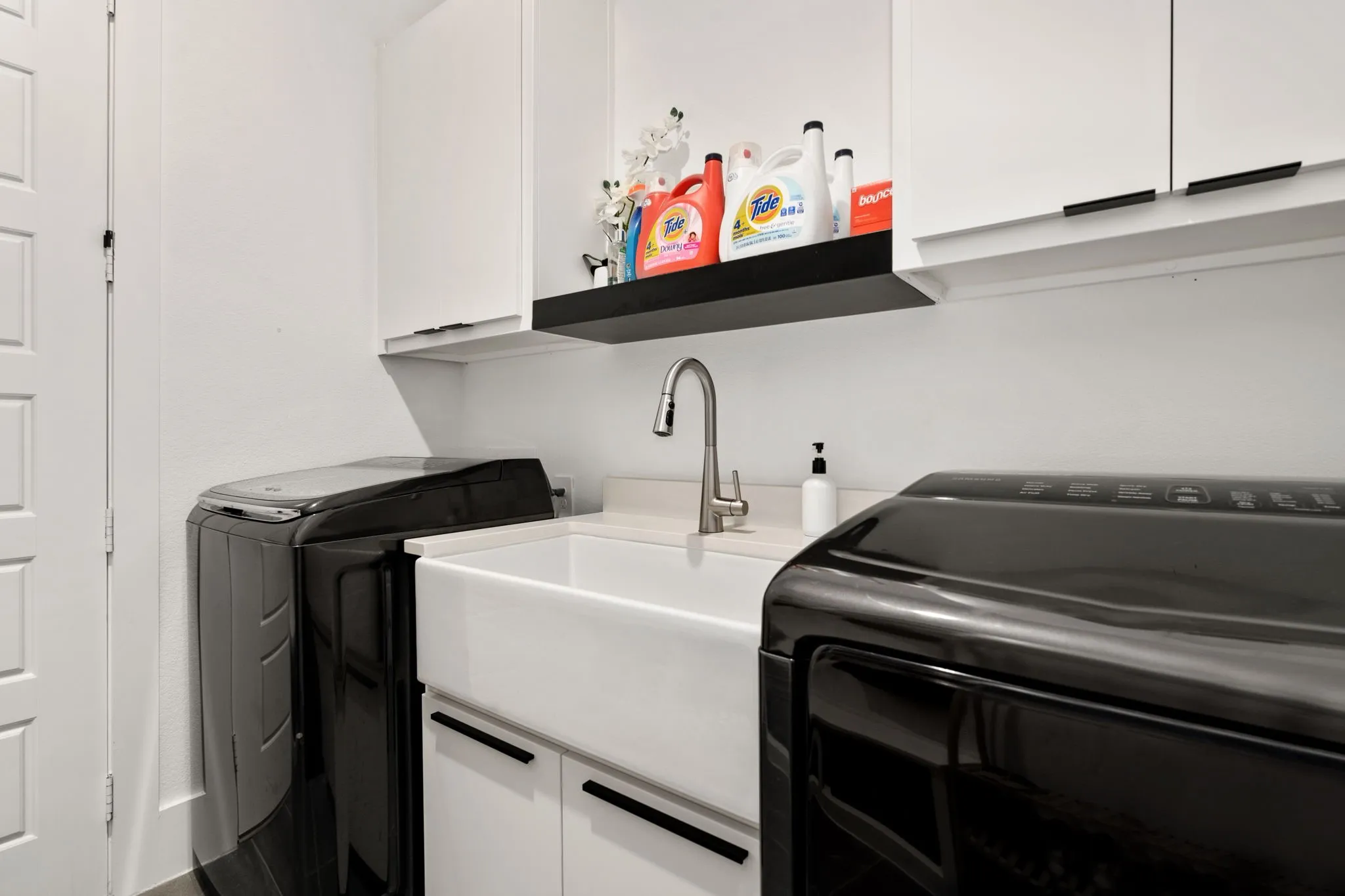 Laundry room featuring a white farmhouse sink with a gooseneck faucet, white cabinetry with black hardware, and an open black floating shelf