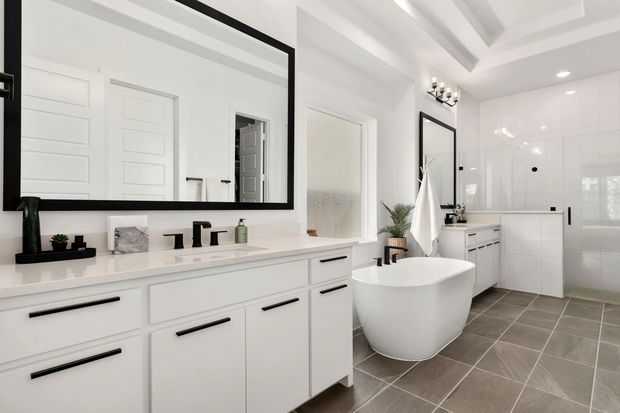 Spacious bathroom featuring a freestanding soaking tub, dual vanities with quartz countertops, a walk-in shower with glass enclosure, white cabinetry, and large format tile flooring