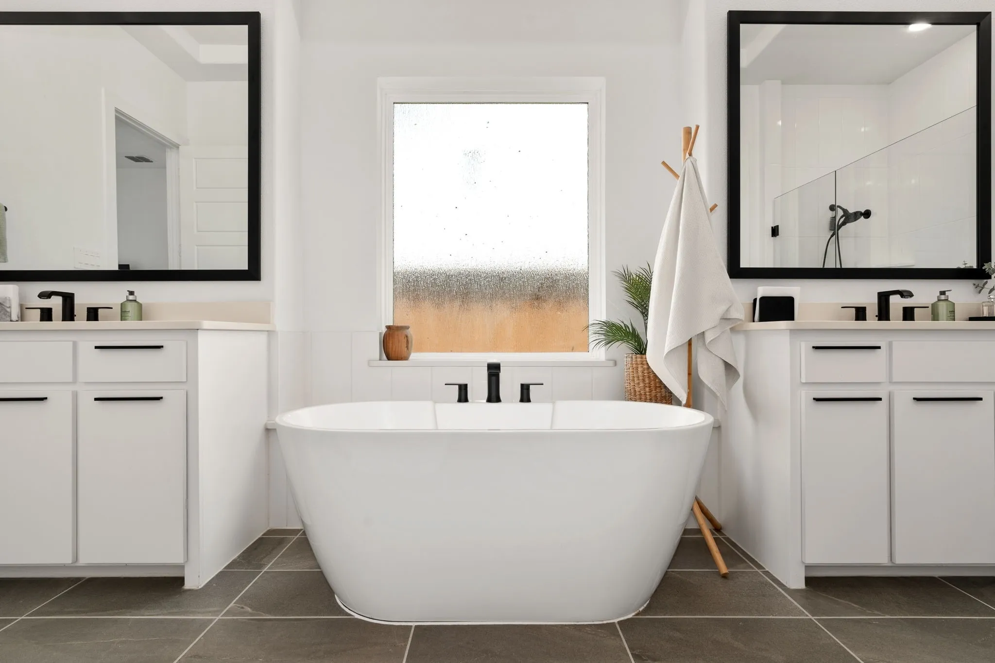 Freestanding soaking tub with dark-finish fixtures, complemented by dual vanities featuring light-tone countertops and under-mount sinks