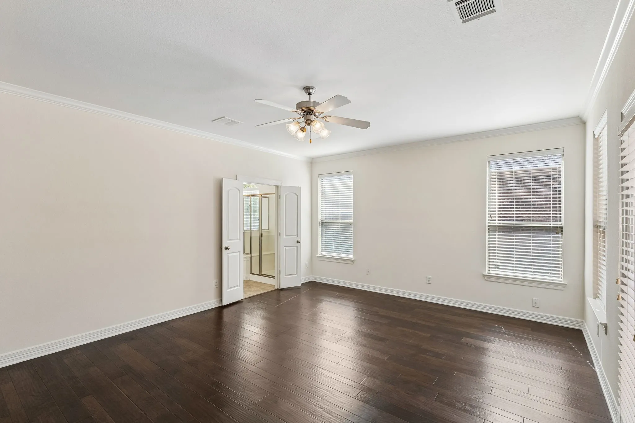 Spacious room featuring dark wood-finish flooring, light neutral walls, a ceiling fan with integrated lighting, and multiple windows with blinds