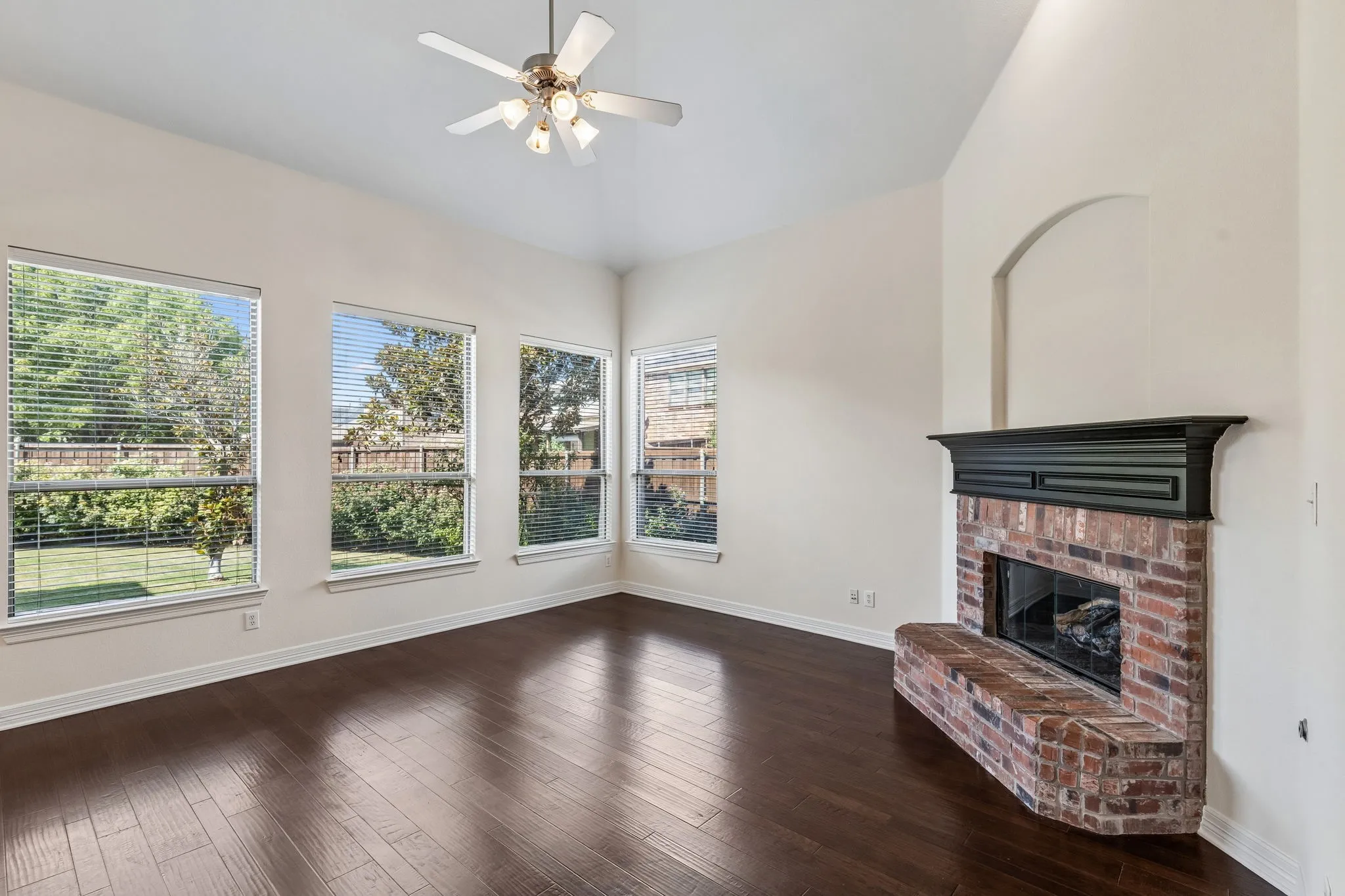 Vaulted ceiling with a ceiling fan, wood-finish flooring, brick fireplace with a dark mantel, multiple windows, and an arched wall niche
