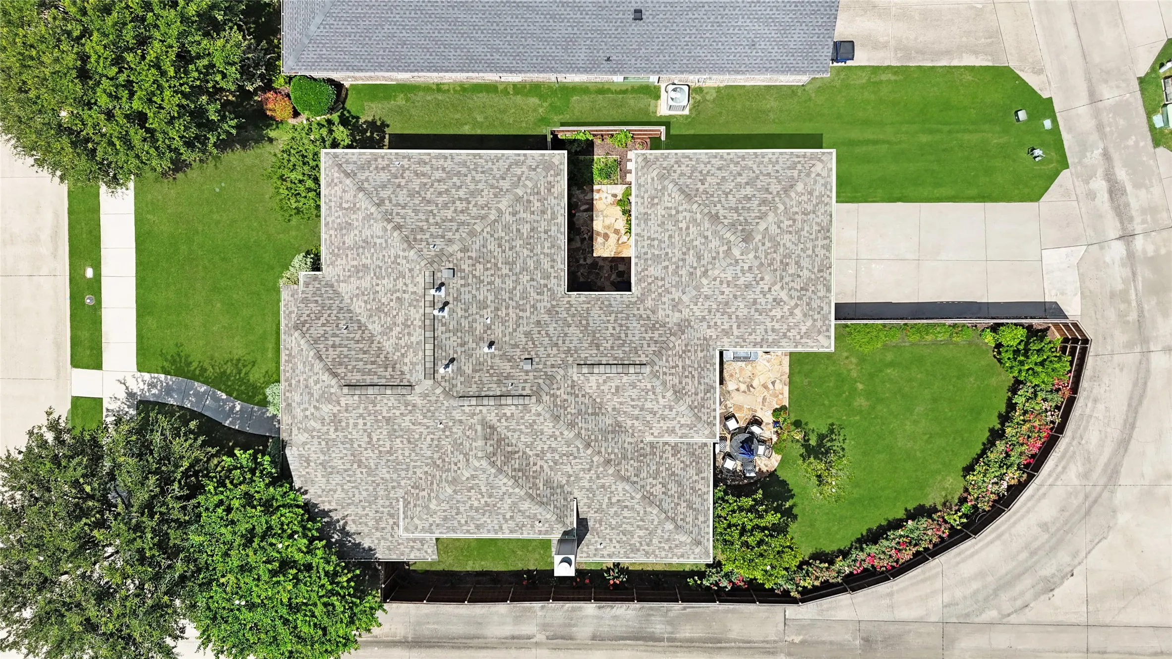 Overhead view of a residential property featuring a shingle roof, paved driveway, concrete walkways, green lawn, and mature trees