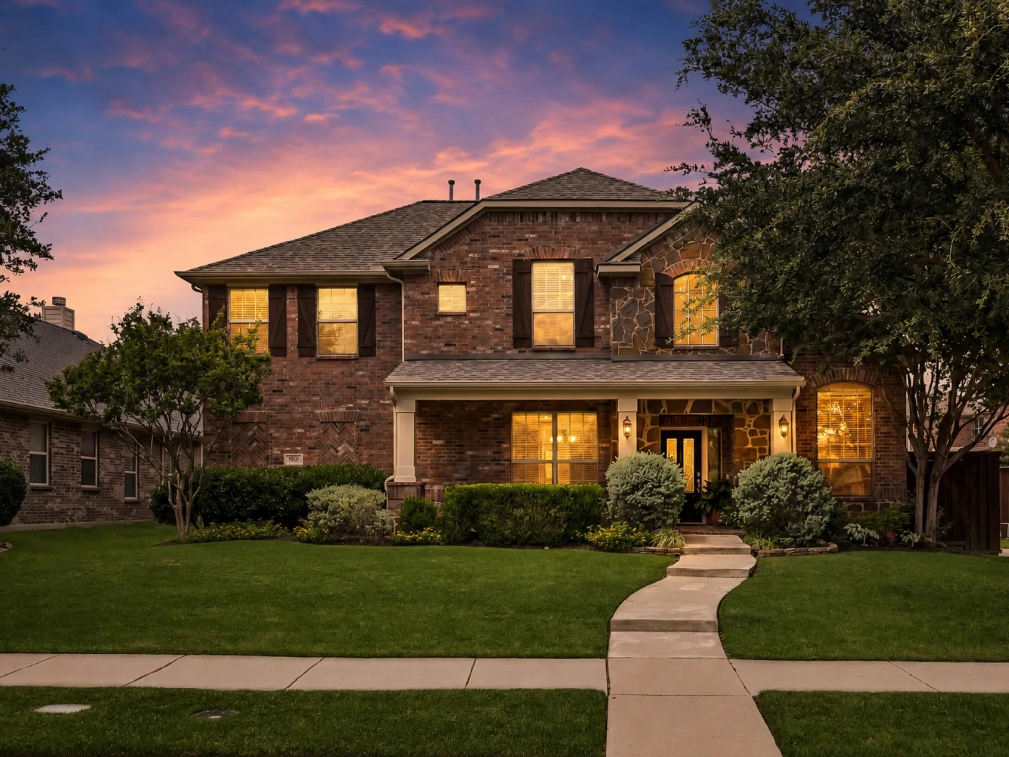 Brick and stone facade residence featuring a covered front porch with columns, a curved concrete walkway, a manicured lawn, and mature landscaping