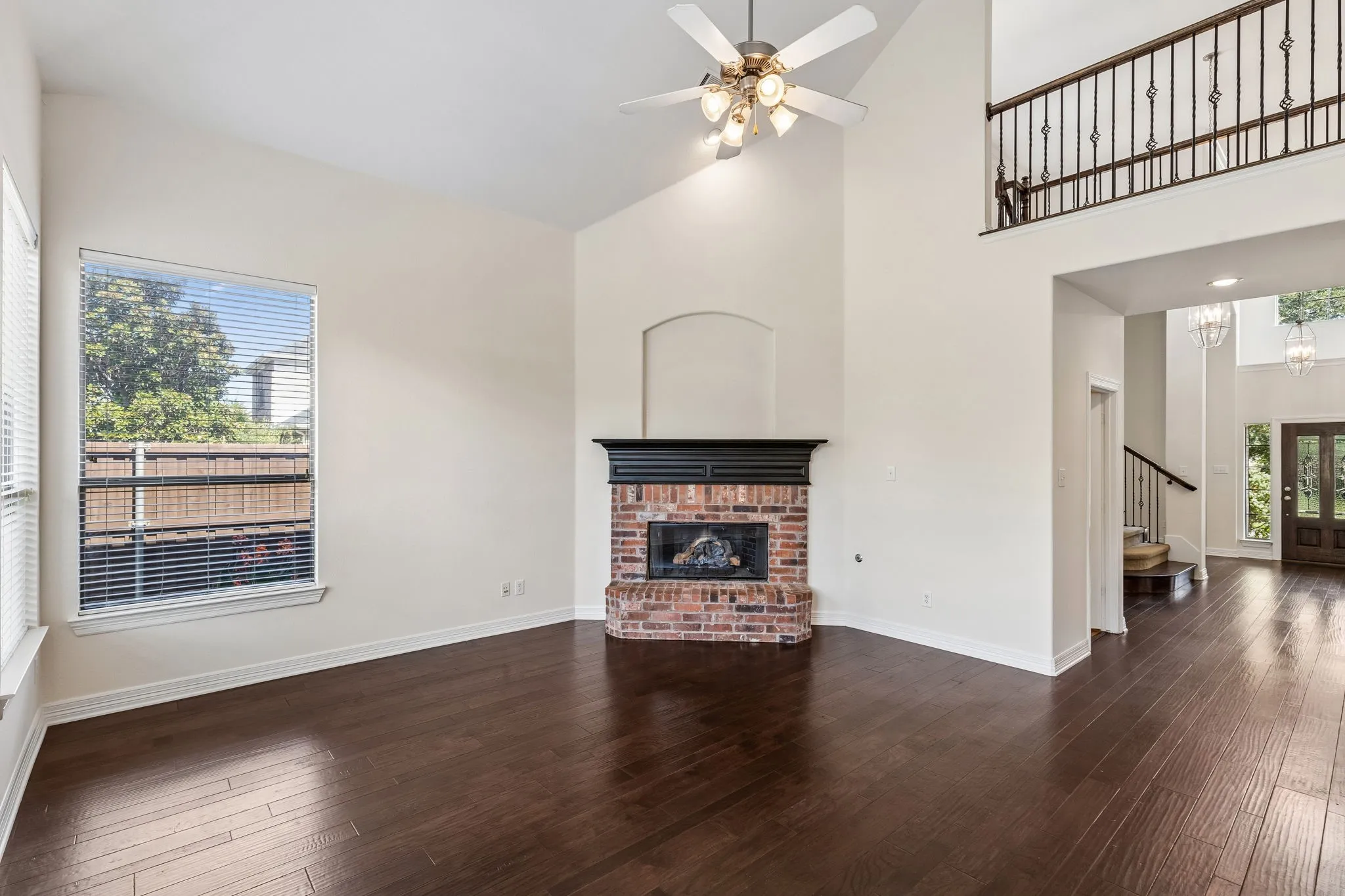 Vaulted ceiling, wood-finish flooring, brick fireplace with mantel, ceiling fan, and an upper-level balcony with metal railings