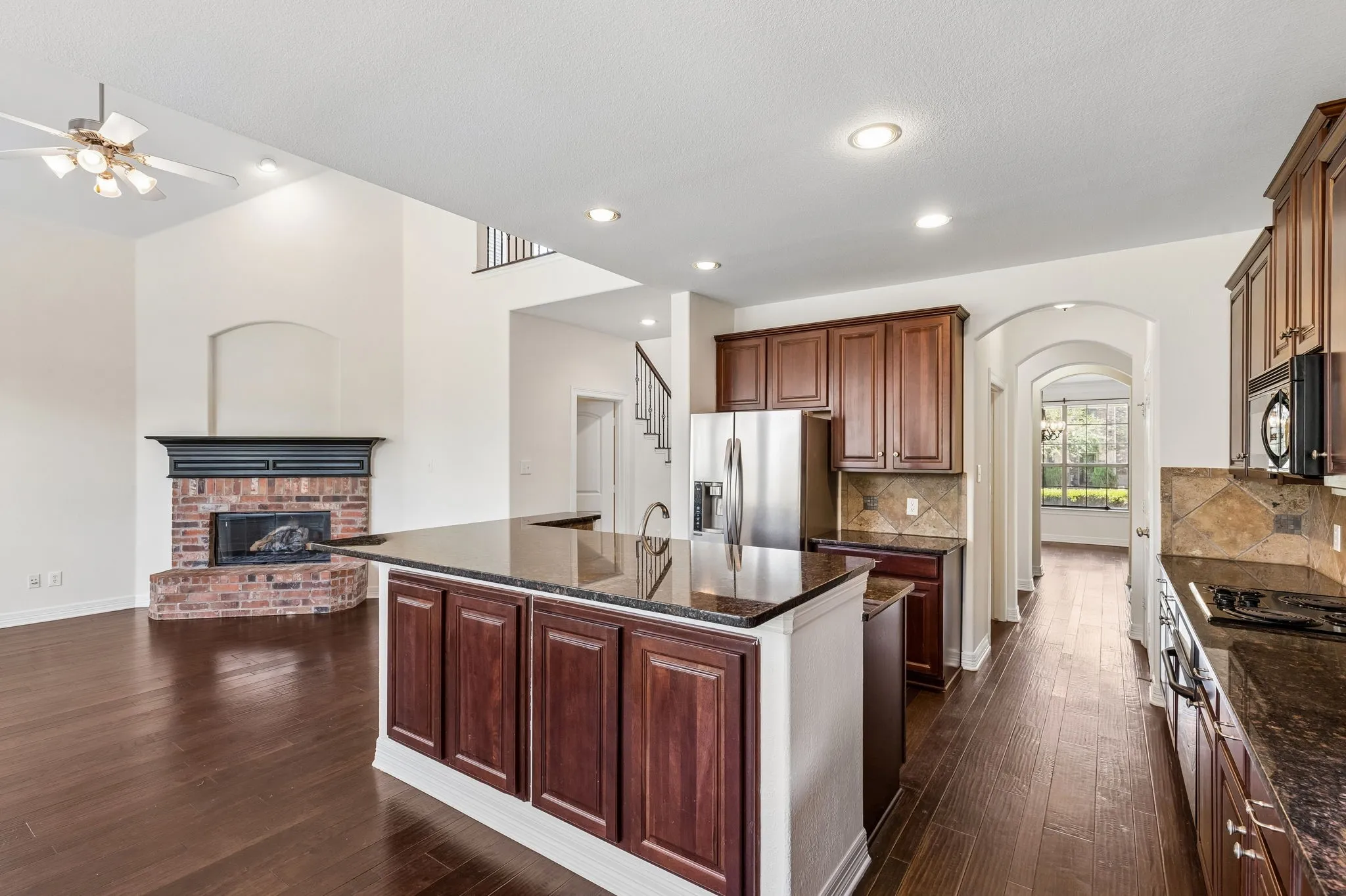 Open-concept kitchen and living area featuring wood-finish flooring, a kitchen island with sink and dark wood cabinetry, stainless steel appliances, and a brick fireplace