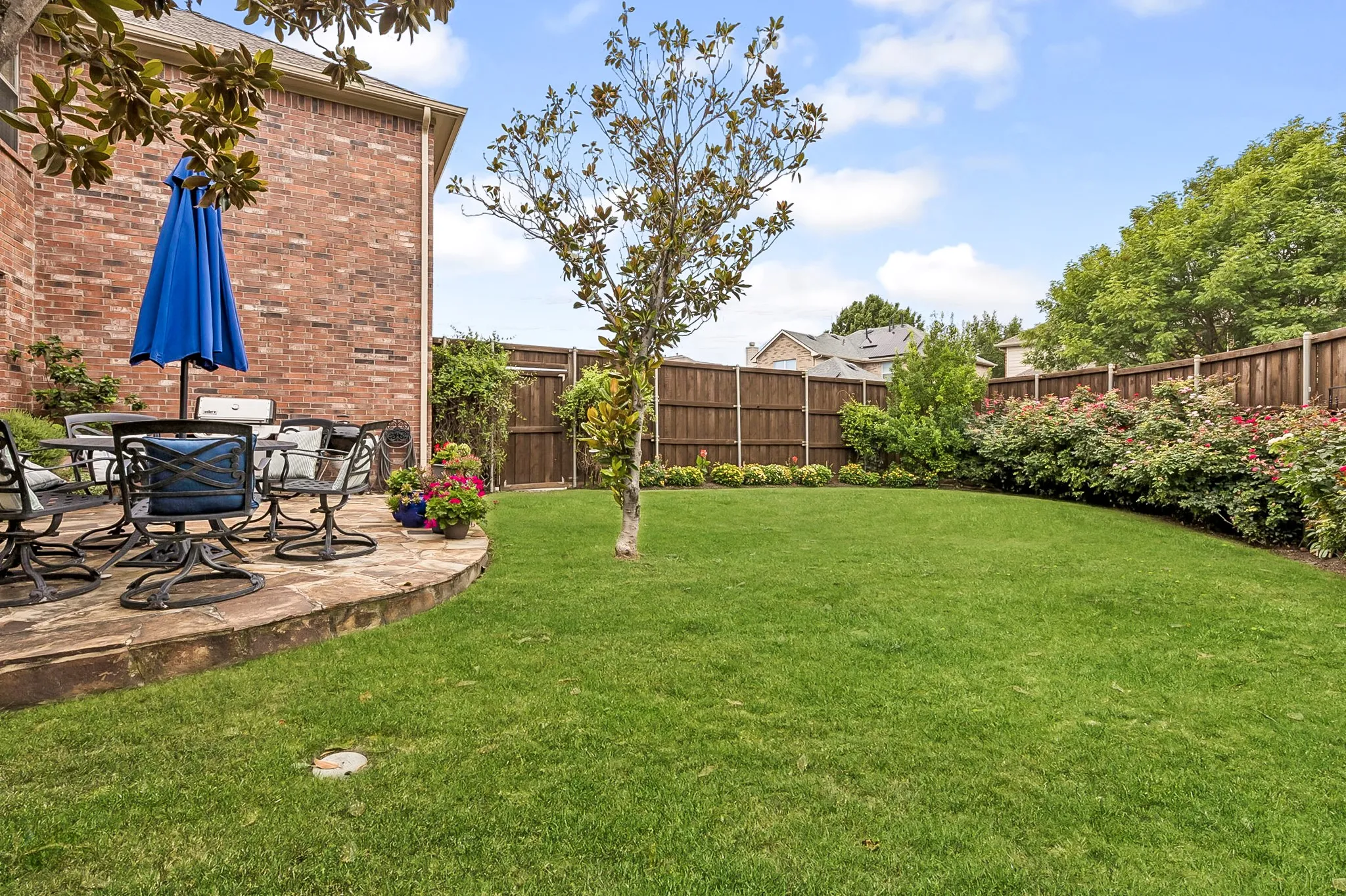 Expansive backyard featuring a stone patio, vibrant green lawn, mature trees, and a perimeter wood fence