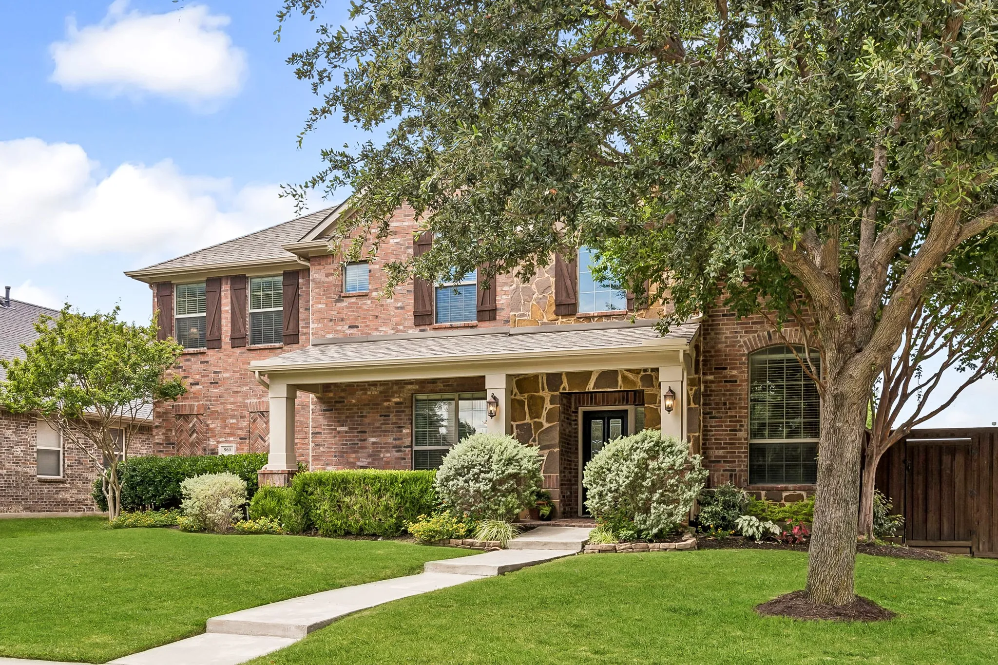 Brick and stone facade residence featuring a covered front porch with columns, a prominent front entry, a manicured lawn, and mature trees