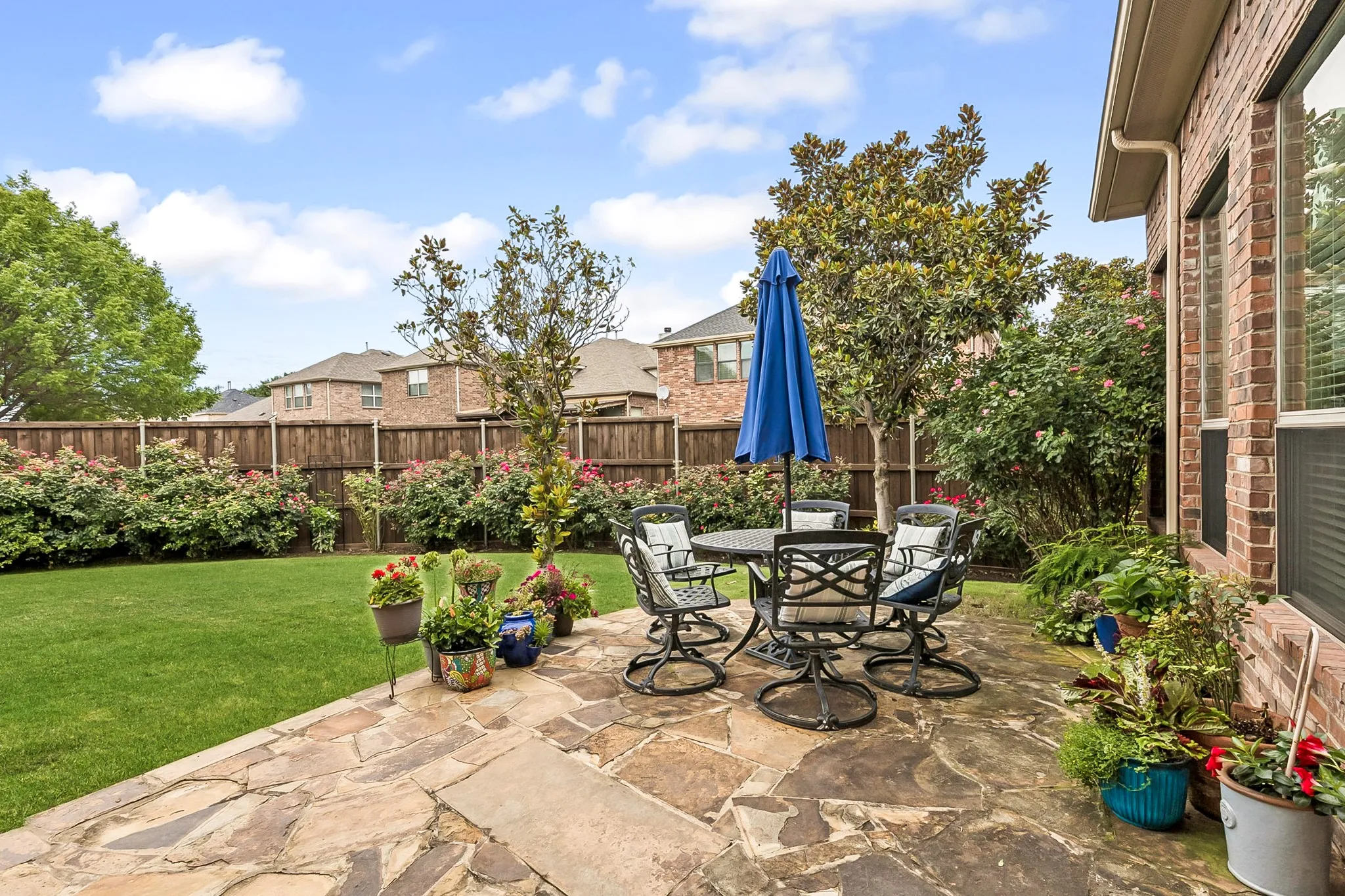 Expansive flagstone patio area featuring a dining set with a blue umbrella, surrounded by lush landscaping, a manicured lawn, and a privacy fence
