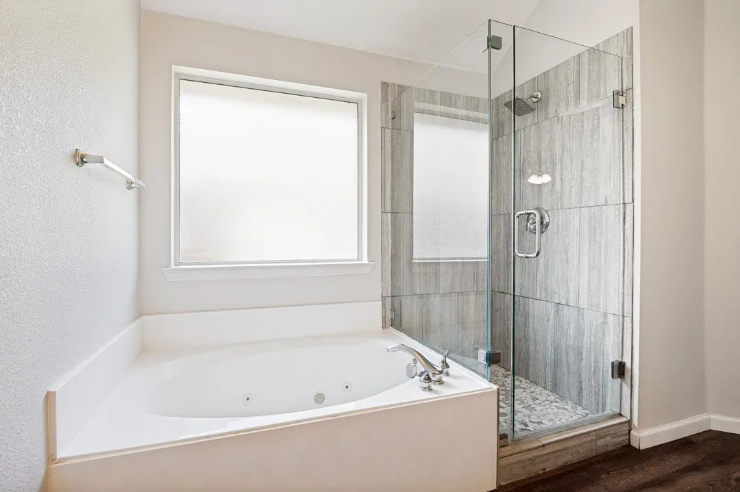 Corner jetted tub with polished chrome fixtures, alongside a glass-enclosed shower featuring stone tile walls, a pebble floor, and a rainfall showerhead