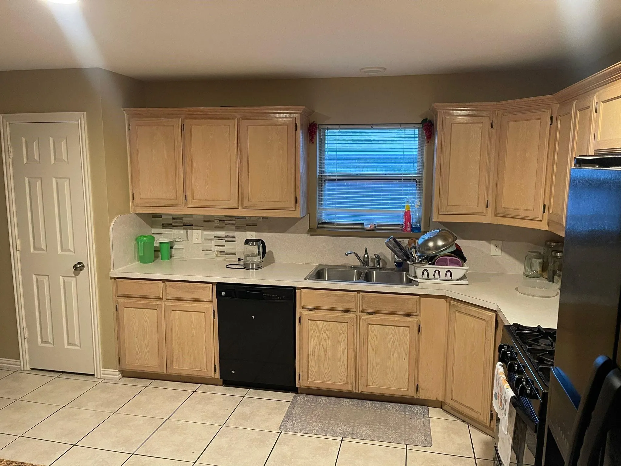 Kitchen featuring light wood-finish cabinetry, a subway tile backsplash, white countertops, a double basin sink, and a black dishwasher