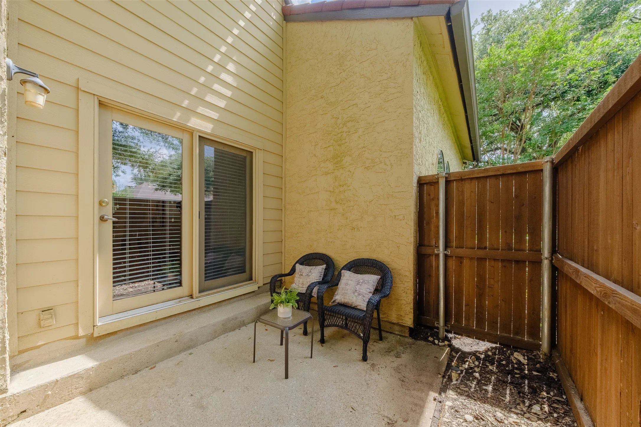 Private fenced back patio with a sliding glass door, wicker seating, and mature tree canopy overhead — a quiet outdoor space tucked away from the street.