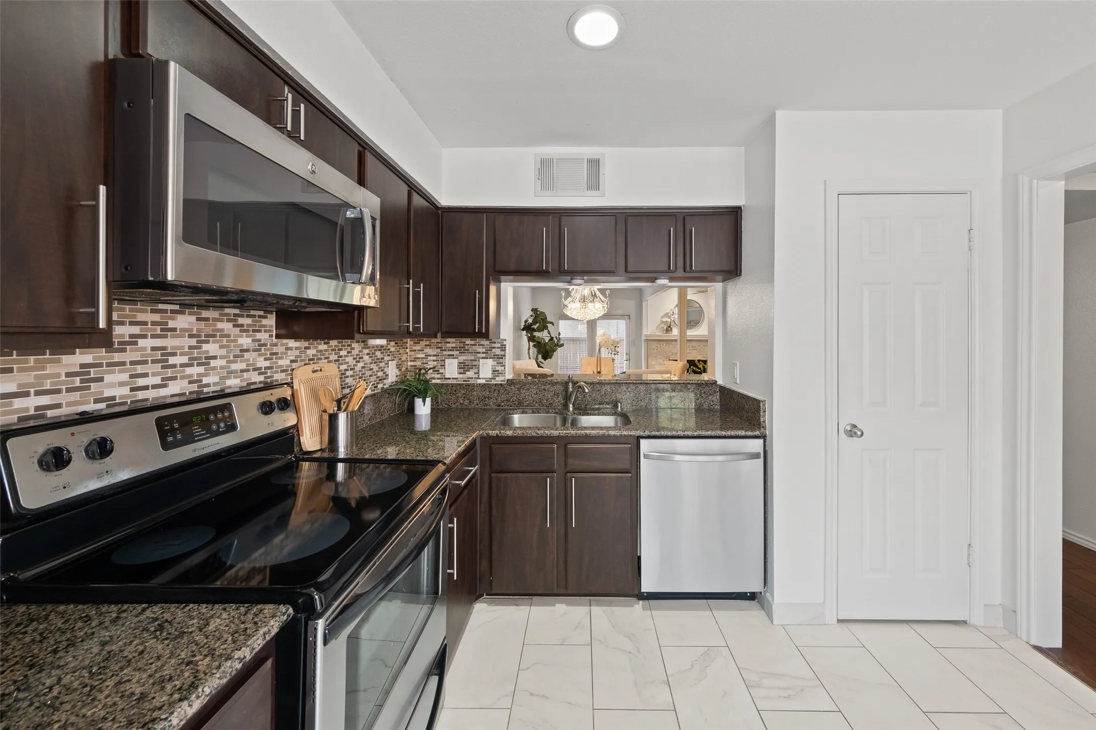 Close-up of the kitchen's stainless steel appliances, granite countertops, mosaic backsplash, and pass-through window framing the dining room chandelier — functional and polished.