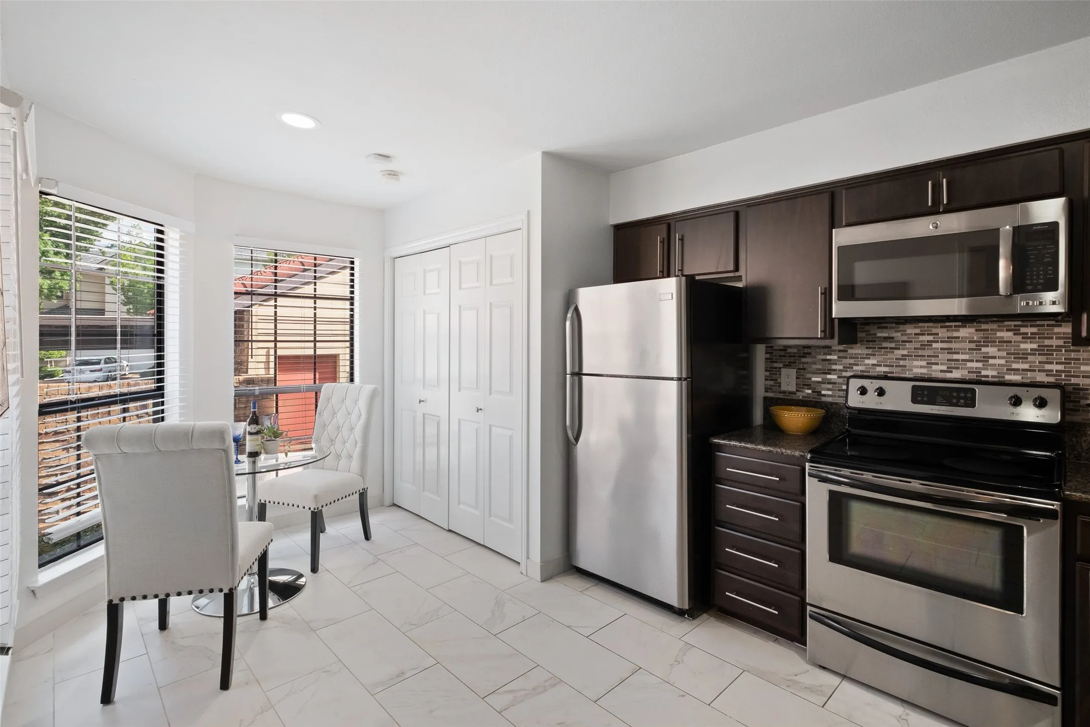 Updated kitchen featuring stainless steel appliances, dark espresso cabinetry, granite countertops, a mosaic tile backsplash, and a sunny breakfast nook with a bistro table and tree views.