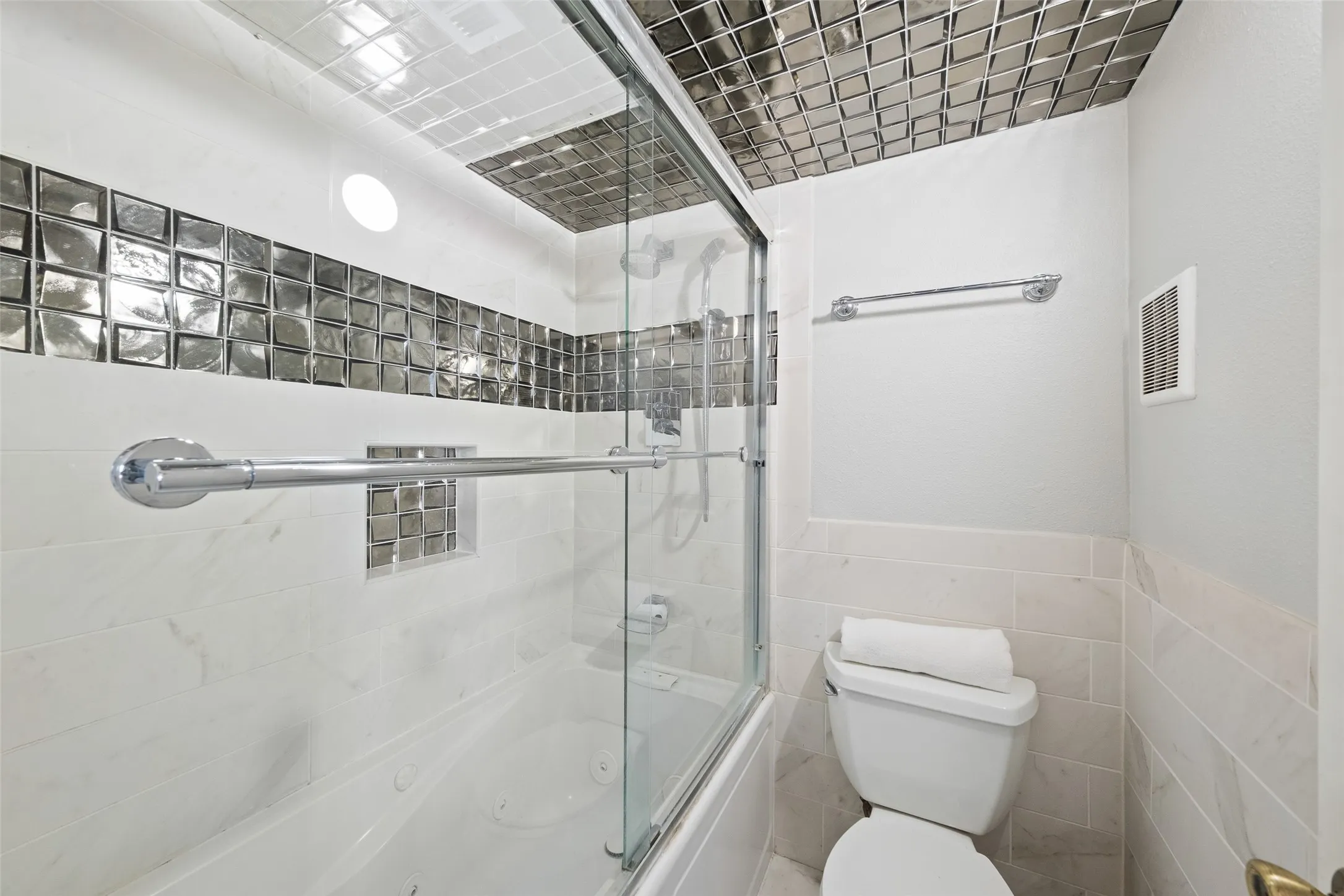 Secondary full bath with a tub/shower combo, glass slider door, reflective mosaic tile accents on the walls and ceiling, grab bar, and a recessed niche — a striking, one-of-a-kind finish.