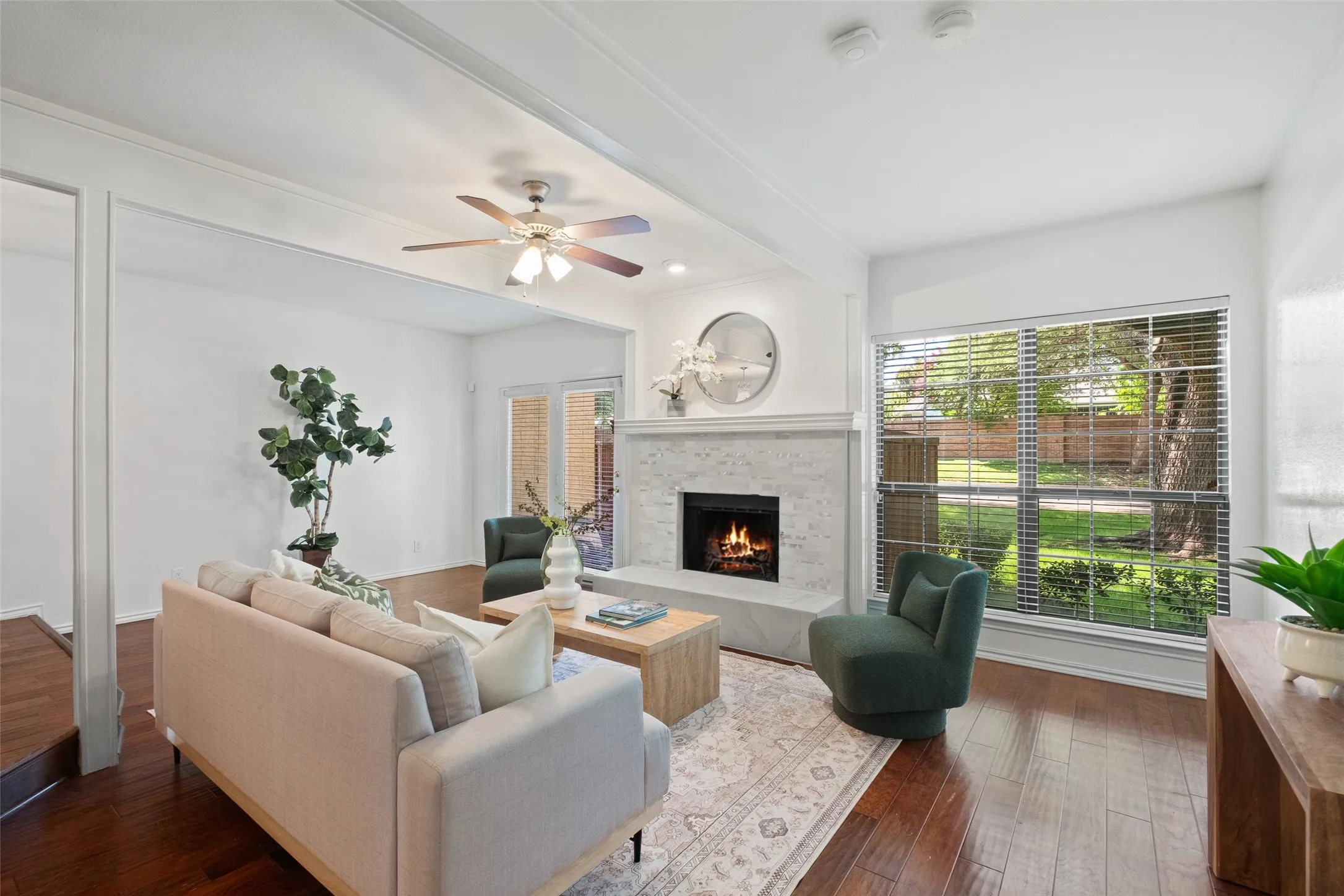 Bright and open living area with hardwood floors, beamed ceiling detail, ceiling fan, and a wall of windows overlooking the lush greenery outside — a rare find in a condo setting.