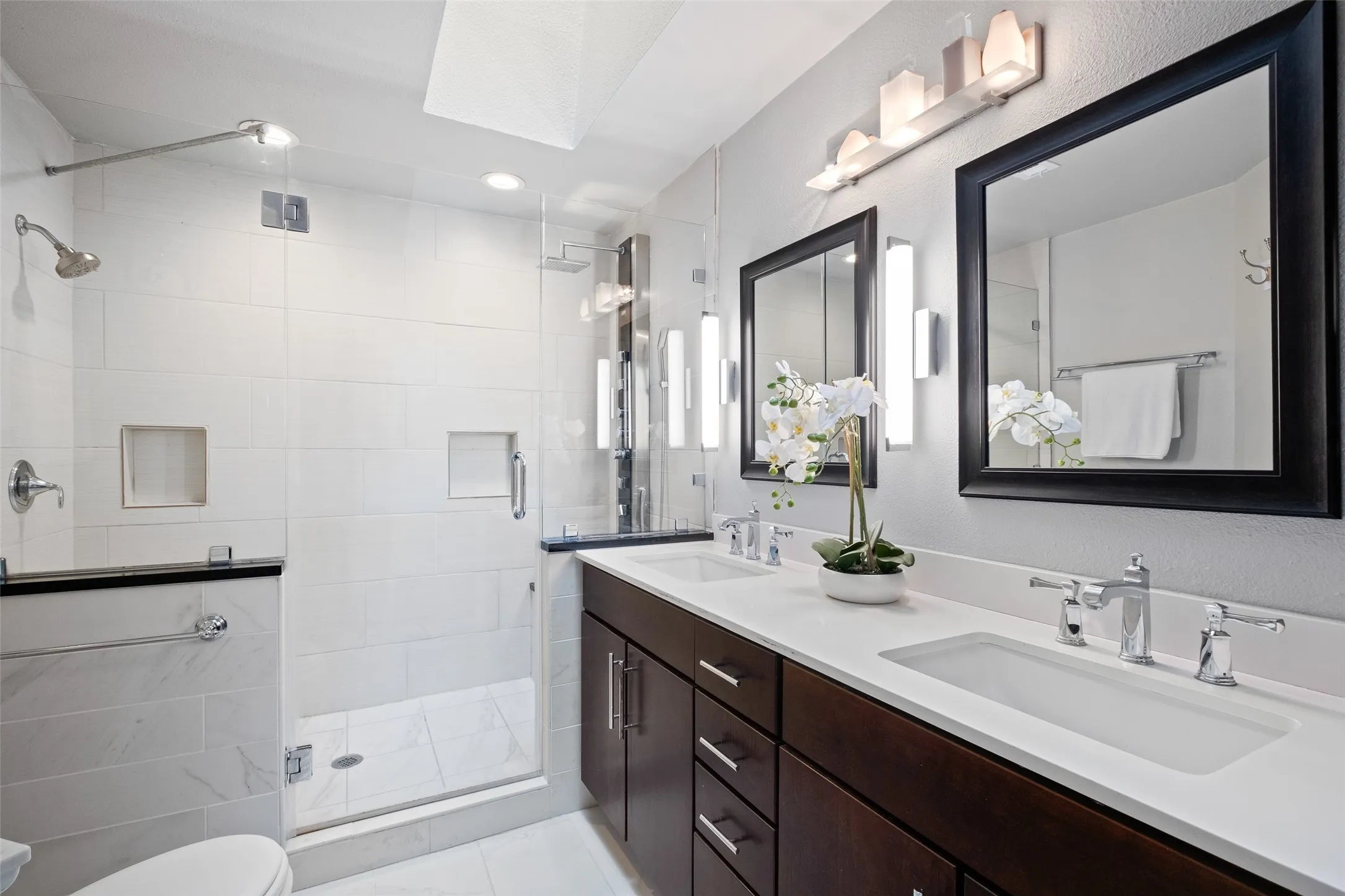 Fully renovated primary bath featuring a double-sink vanity with quartz countertops, dark espresso cabinetry, framed mirrors, modern sconce lighting, and a frameless glass walk-in shower with large-format white tile.