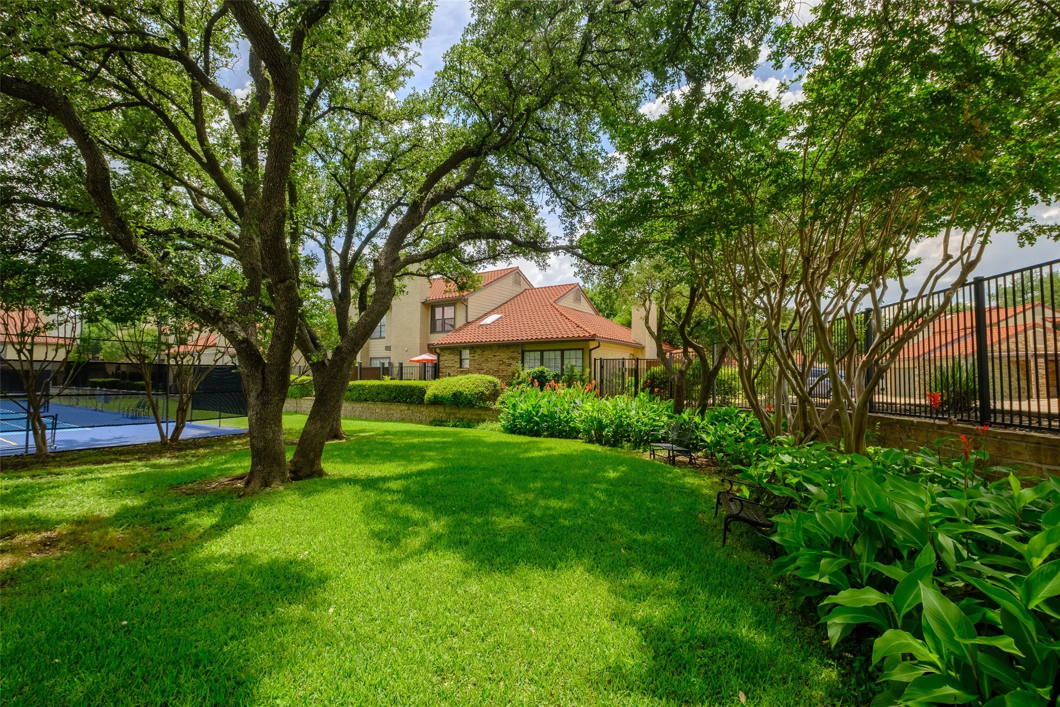 Beautifully landscaped community common area with sweeping lawn, mature live oaks, colorful flowering beds, and a tucked-away bench
