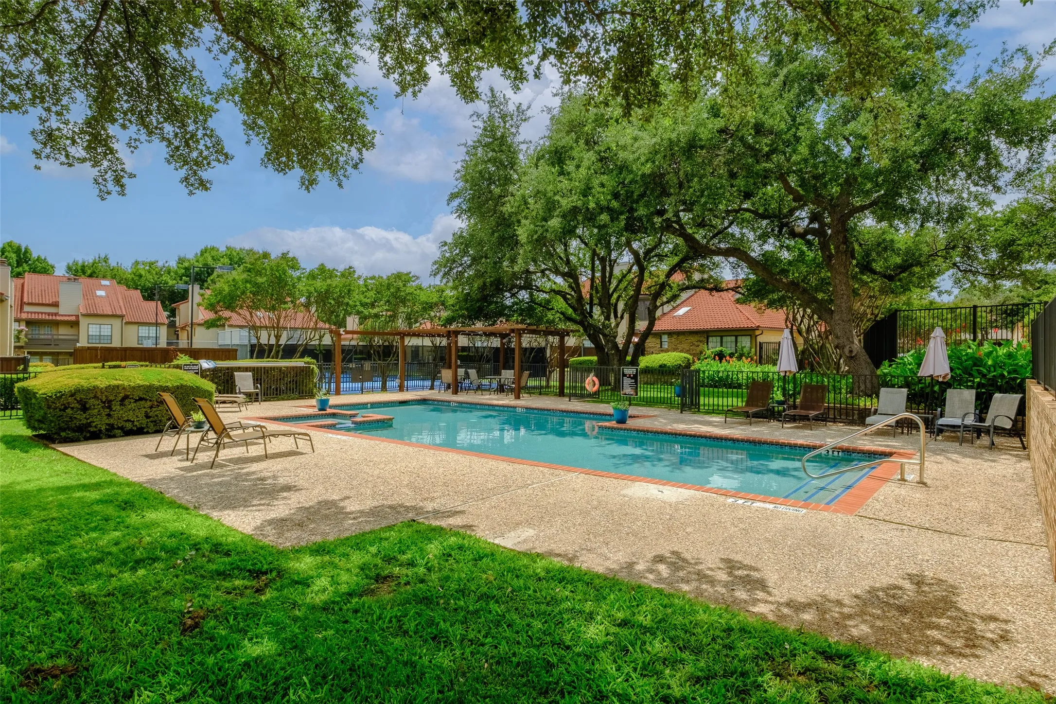 Lush, tree-shaded view of the community pool and sun deck with lounge seating, a pergola-covered gathering area, and the La Mirada roofline as a backdrop — the definition of low-maintenance resort living.