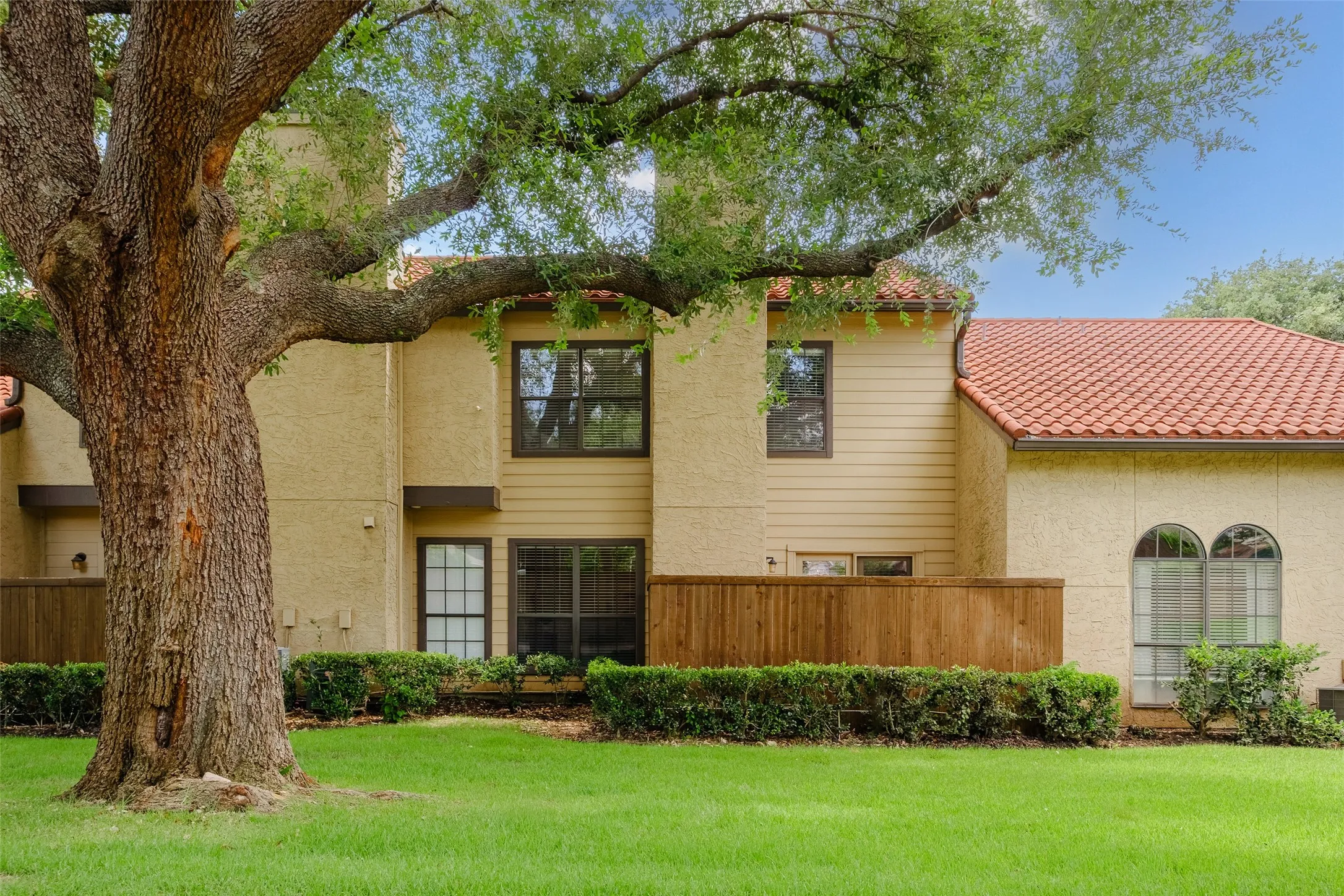 Back exterior view showcasing the clay tile roof, private wood fence patio enclosure, mature live oak canopy, and lush community greenspace.