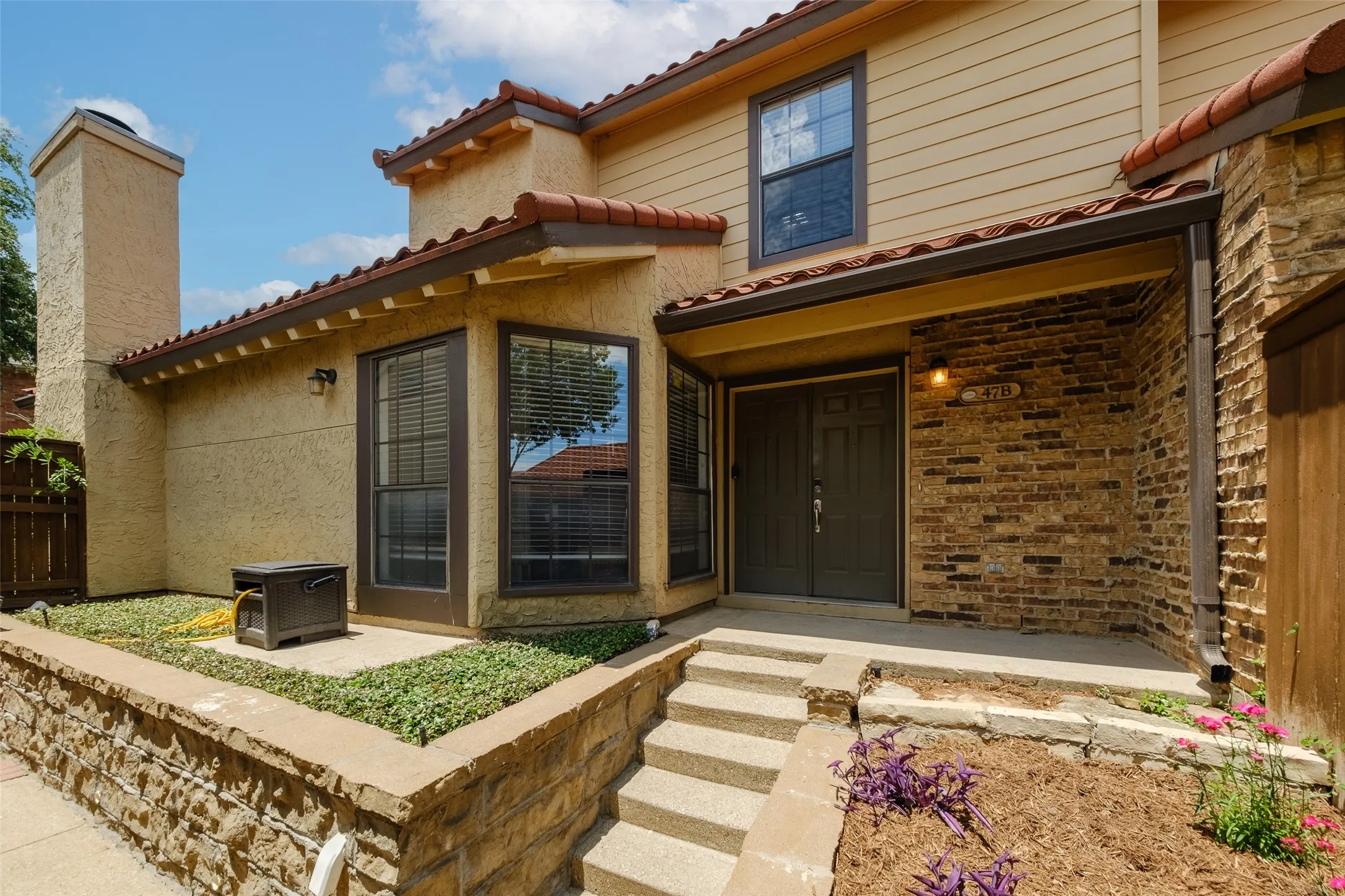 Close-up of the covered front entry with brick surround, tile roof overhang, unit signage, and blooming landscape plantings framing the steps — a welcoming and private arrival experience.