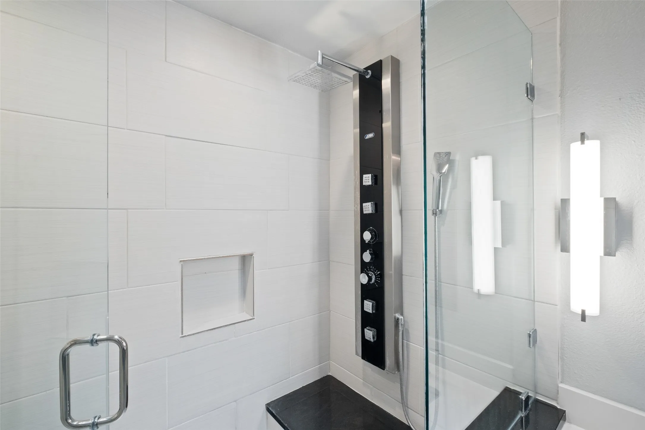 Walk-in shower with a full multi-function shower panel system, rainfall head, built-in niche, marble-look floor tile, and frameless glass enclosure with bench seating.