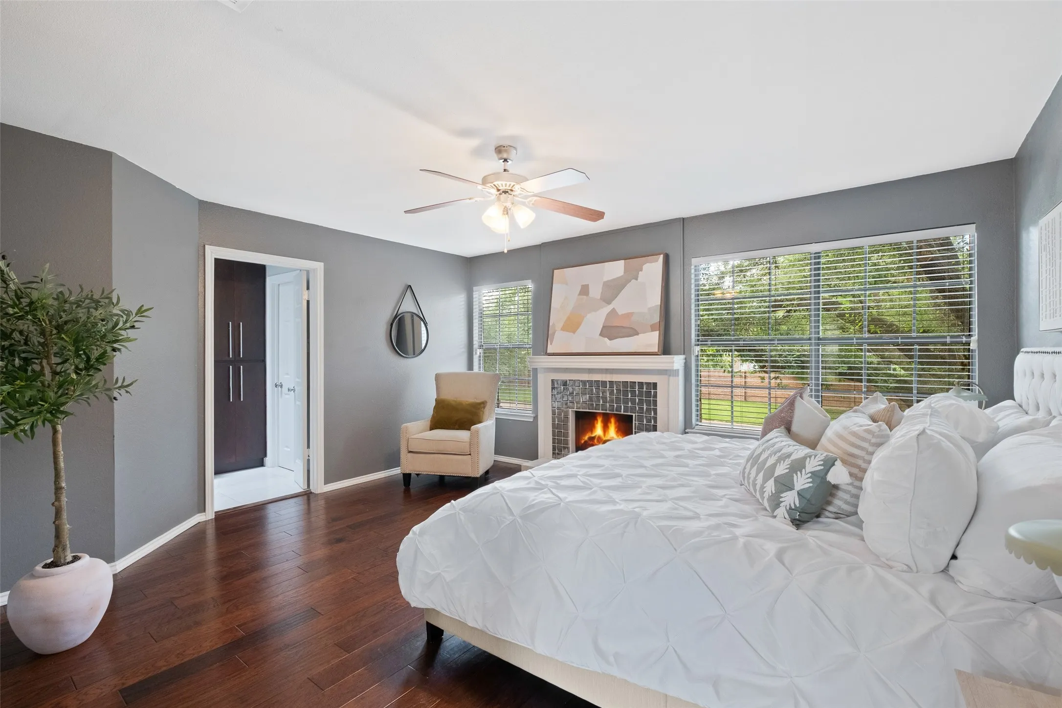 Spacious primary bedroom with hardwood floors, a second wood-burning fireplace with tile surround, multiple windows overlooking the greenery, ceiling fan, and direct access to the primary bath and walk-in closet.