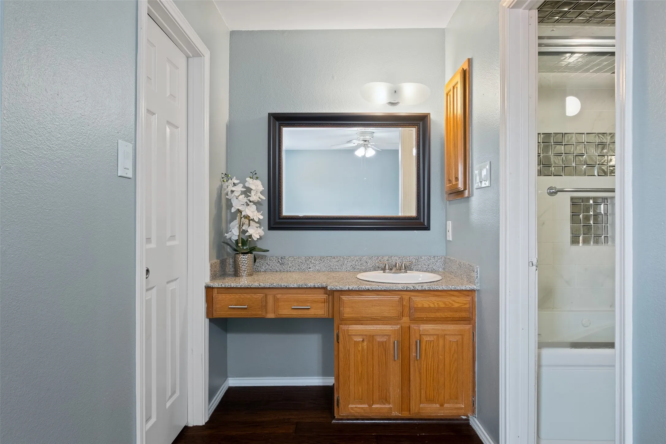 Secondary bathroom vanity area with granite countertop, warm wood cabinetry, a large framed mirror, and a peek into the shower/tub combo beyond.
