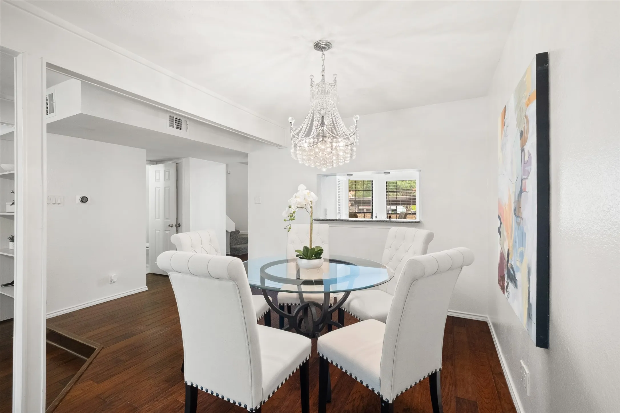 Elegant dining room with a statement crystal chandelier, glass-top table, hardwood floors, and open sight lines into the living area — a natural gathering space for dinner parties.