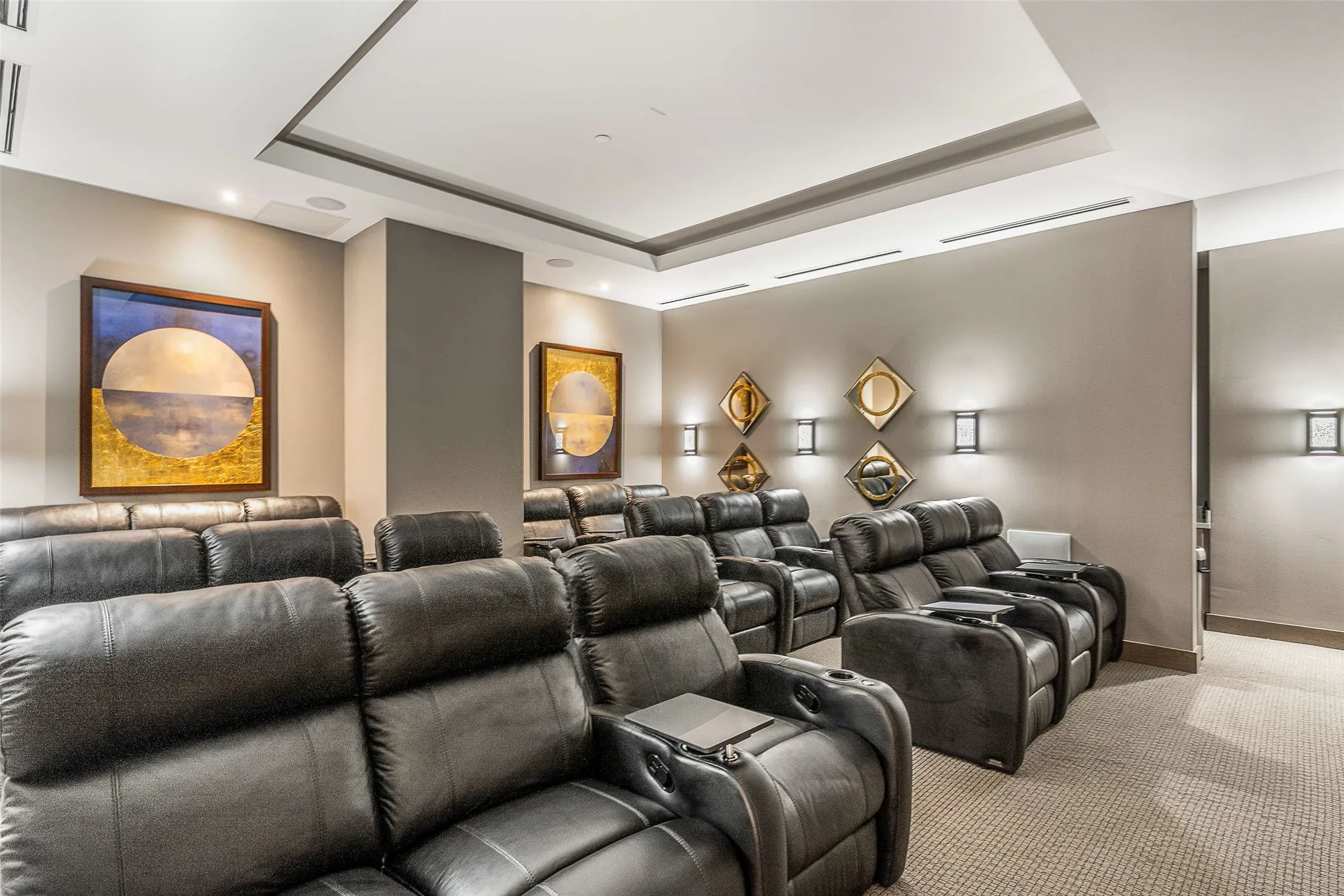 Community Dedicated media room featuring tiered seating with multiple rows of dark upholstered recliners, wall-mounted sconce lighting, integrated ceiling speakers, recessed lighting, and a tray ceiling with accent lighting