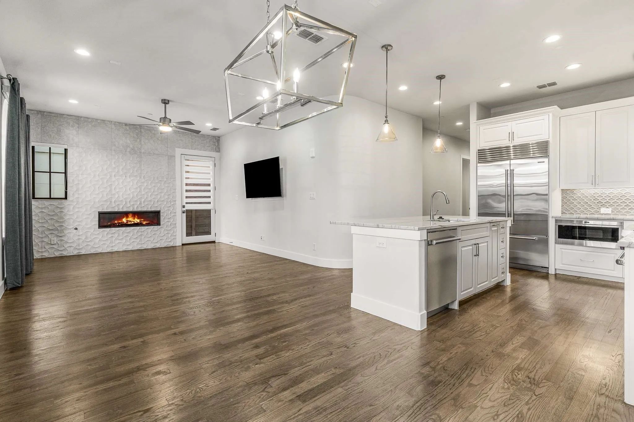 Open-concept living space featuring wood-finish flooring, a contemporary fireplace with textured wall surround, white cabinetry, a kitchen island with sink, and stainless steel appliances